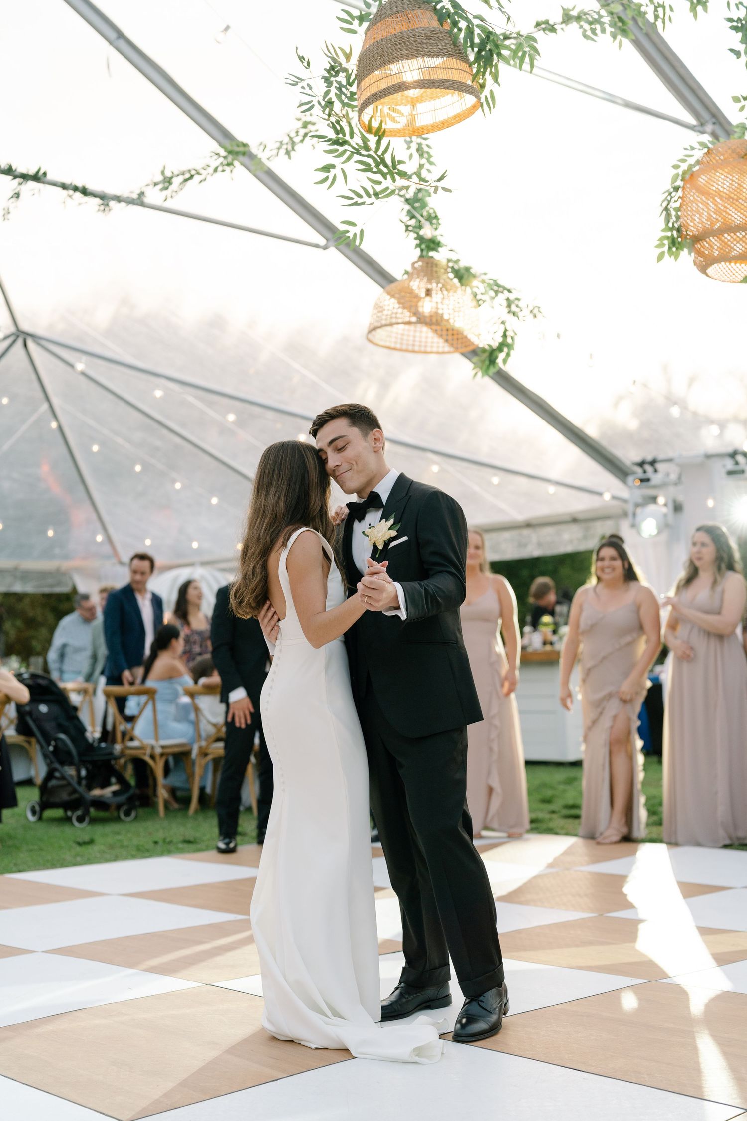 Bride and groom share their first dance under a clear tent with hanging rattan lights.