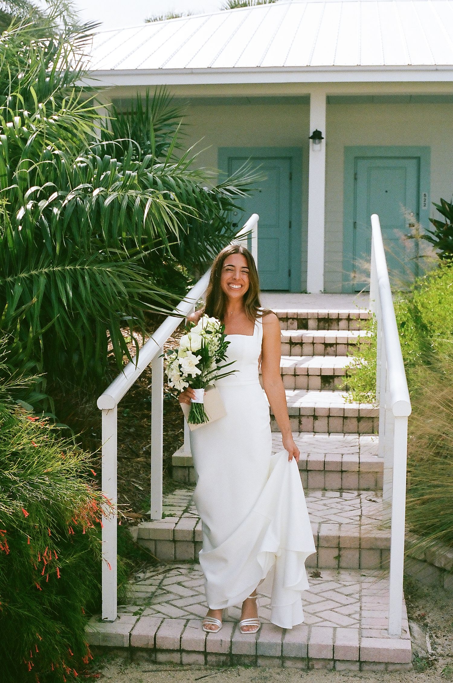 Smiling bride in white gown holding bouquet on steps of a charming turquoise cottage surrounded by tropical greenery.