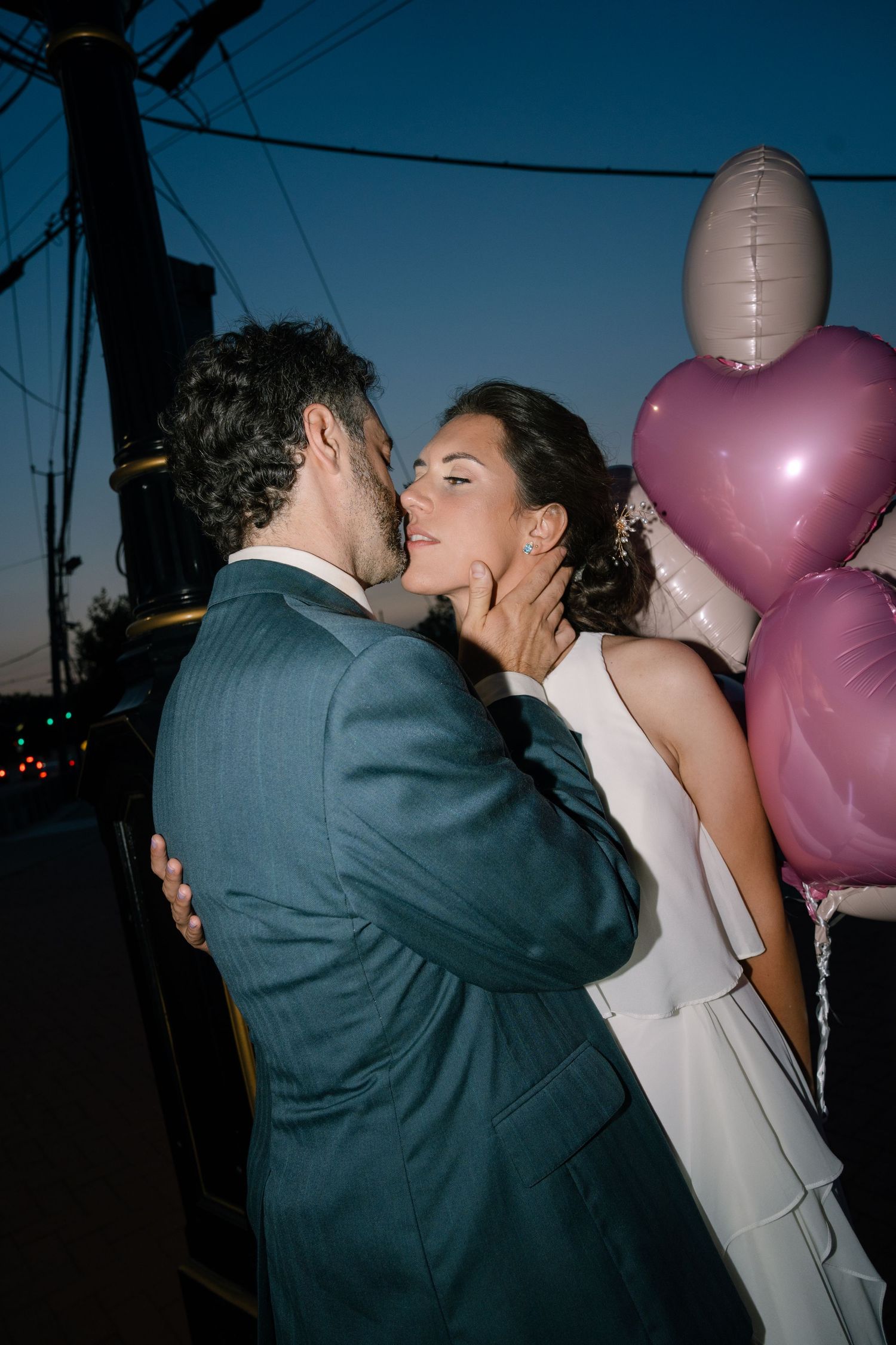 A couple sharing a romantic kiss outdoors at dusk with pink balloons floating in the background.