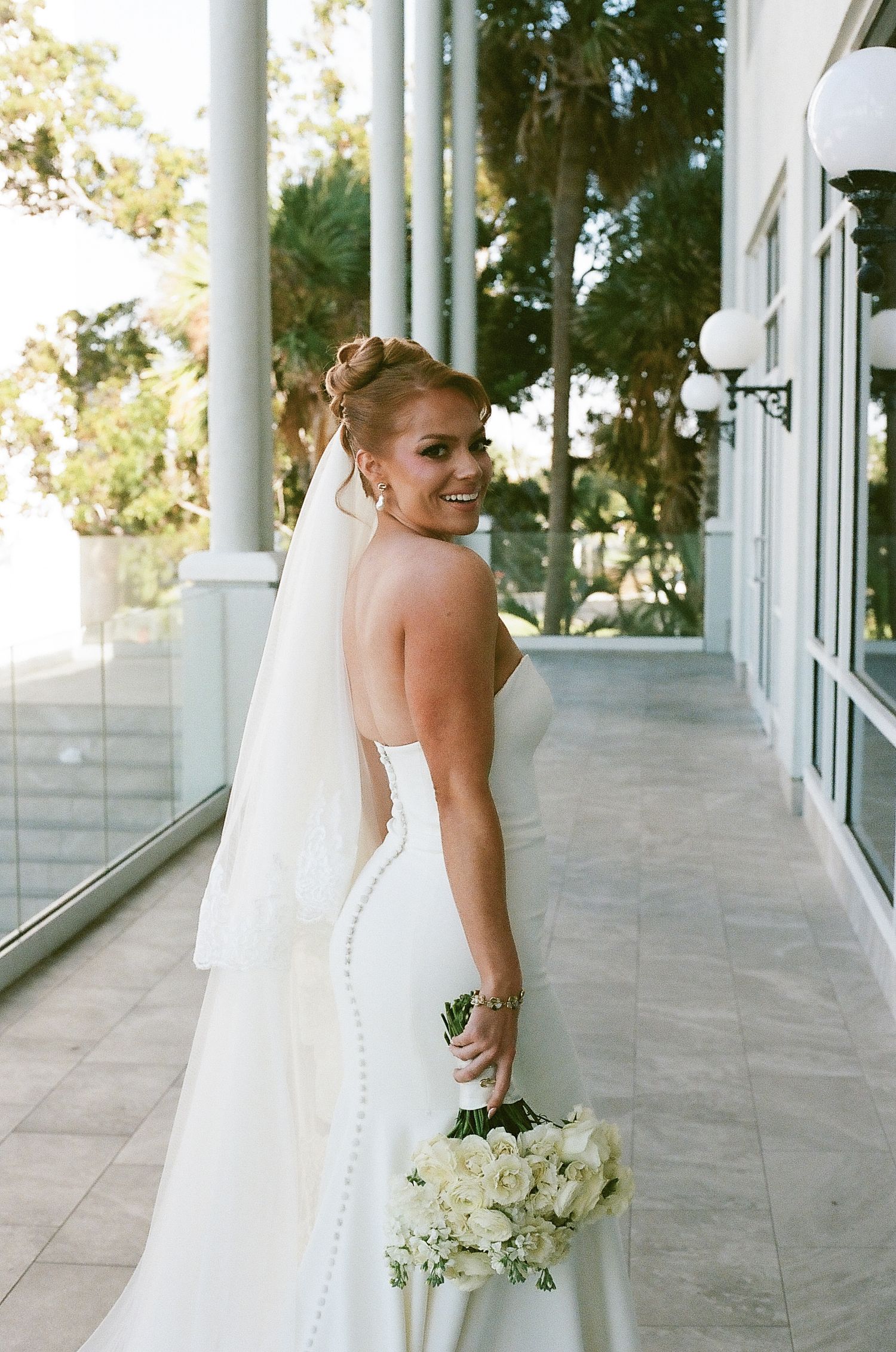 A bride in a white wedding gown with buttons down the back stands in an elegant columned hallway holding a bouquet.