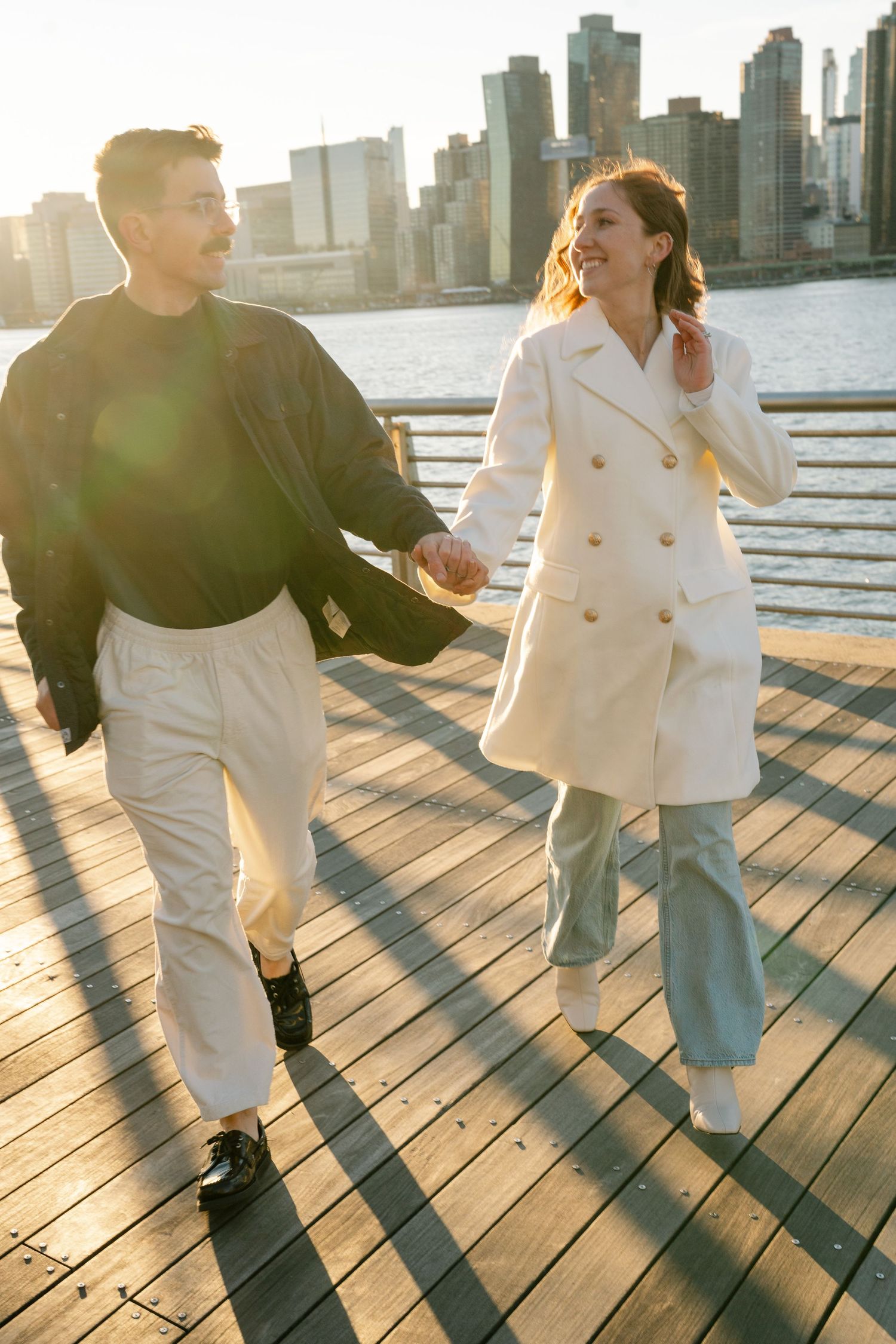 A joyful couple walks along a wooden pier during golden hour with the Manhattan skyline in view.