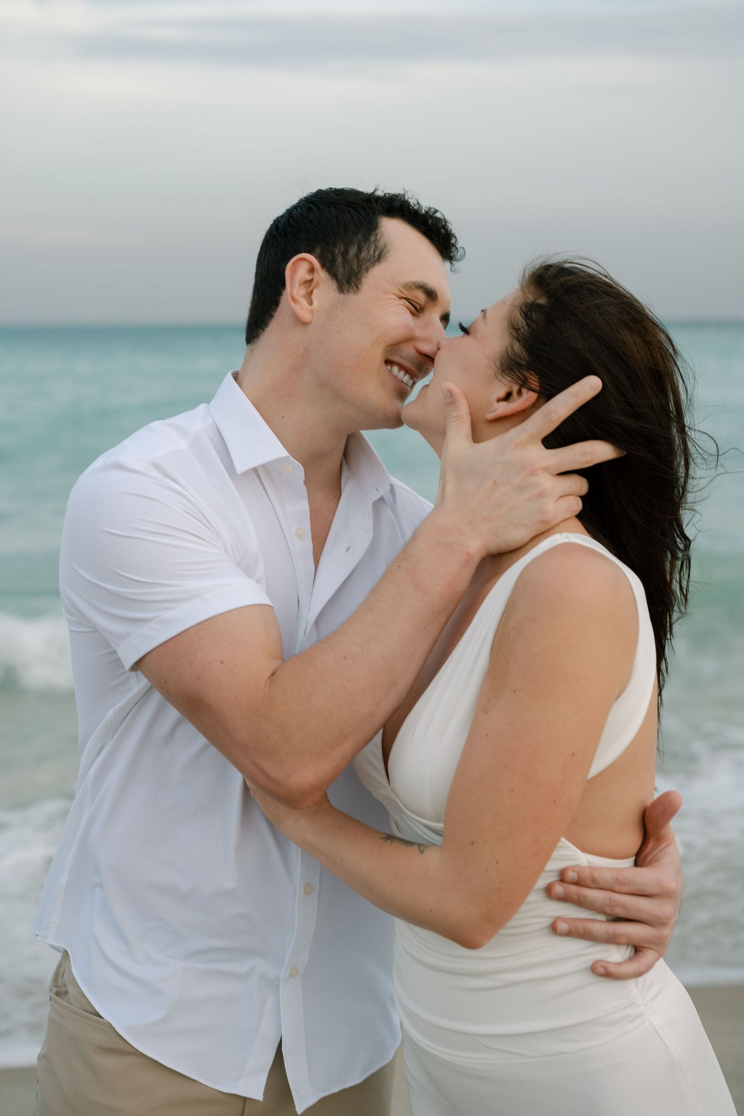 A couple shares intimate moments on the beach wearing white and beige outfits against an ocean backdrop.