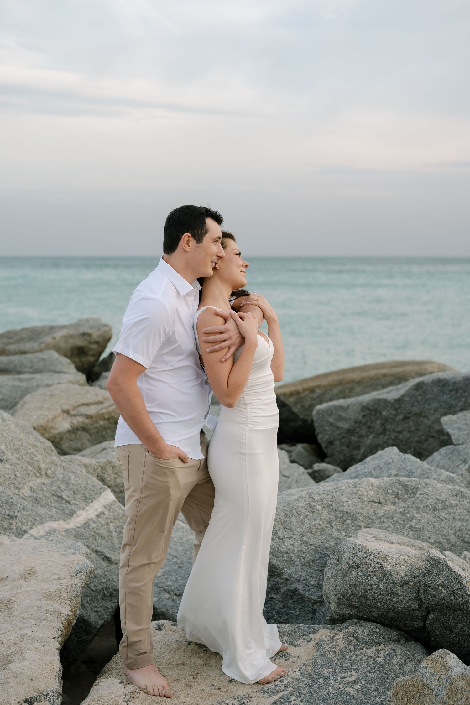 A couple in white attire embraces on rocky beach terrain during sunset with turquoise ocean waters in background.