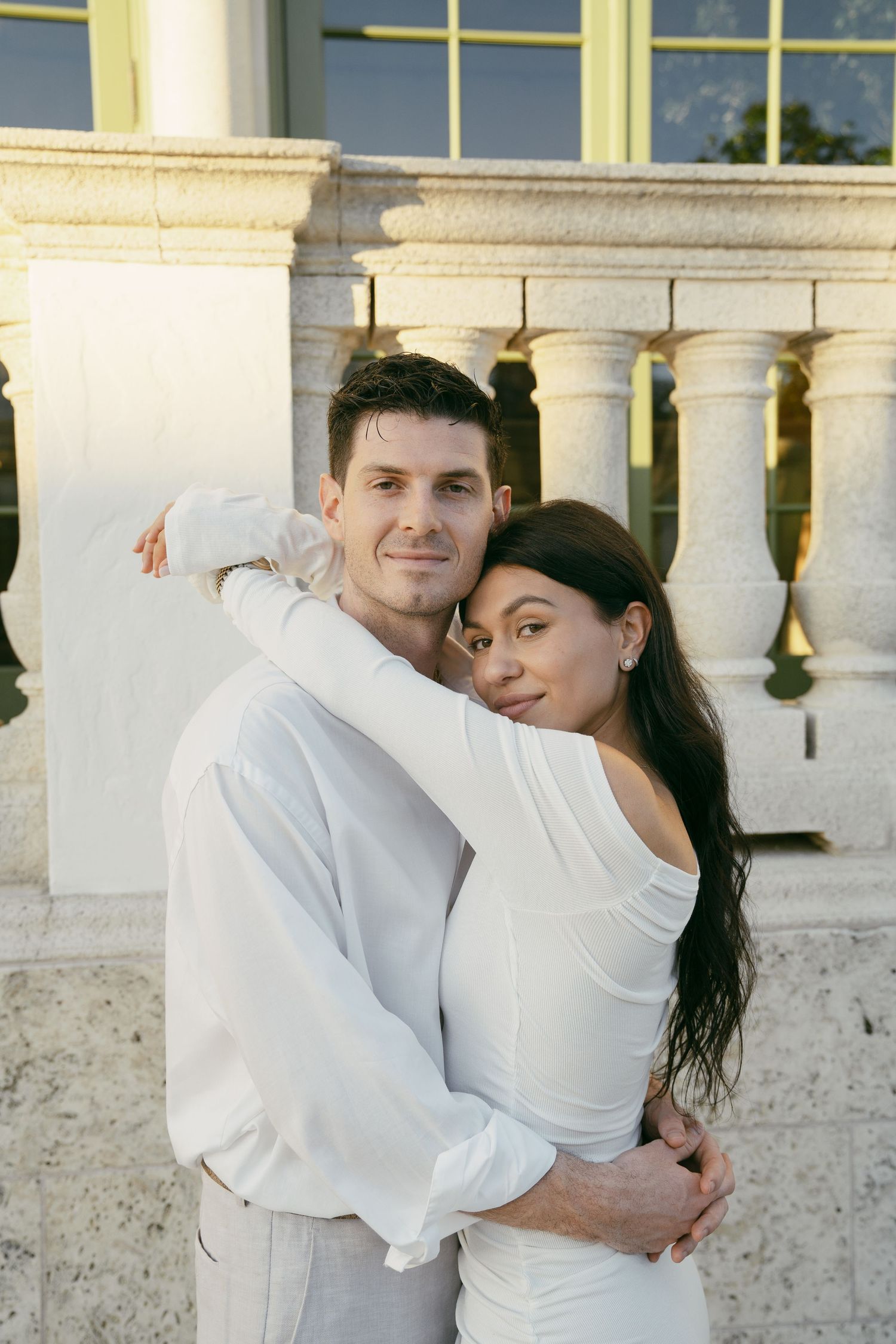 A loving couple in white attire embraces affectionately against a white columned architectural backdrop in a romantic scene.