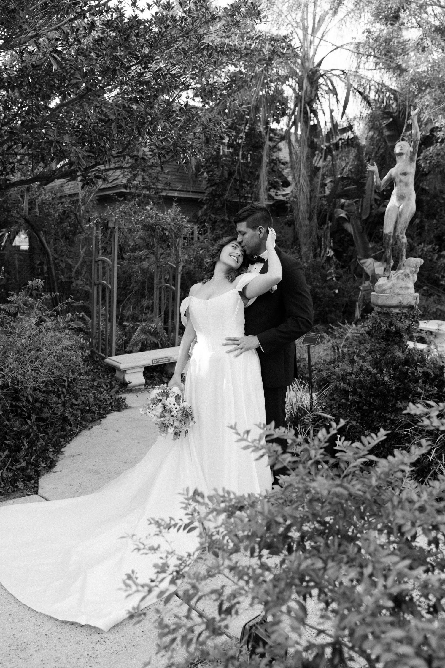 A black and white wedding photograph in a lush garden setting with a long flowing white dress and floral bouquet.