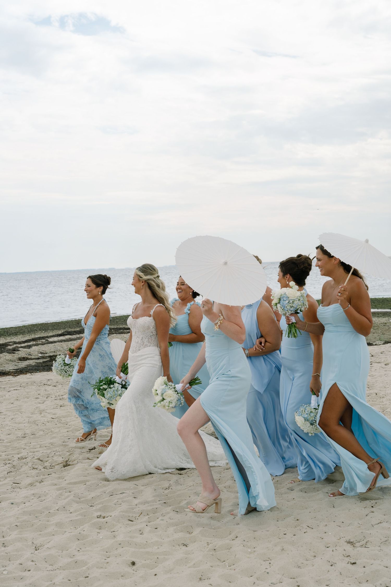 Bride and bridesmaids in light blue gowns walk on a beach, some holding white parasols and bouquets.