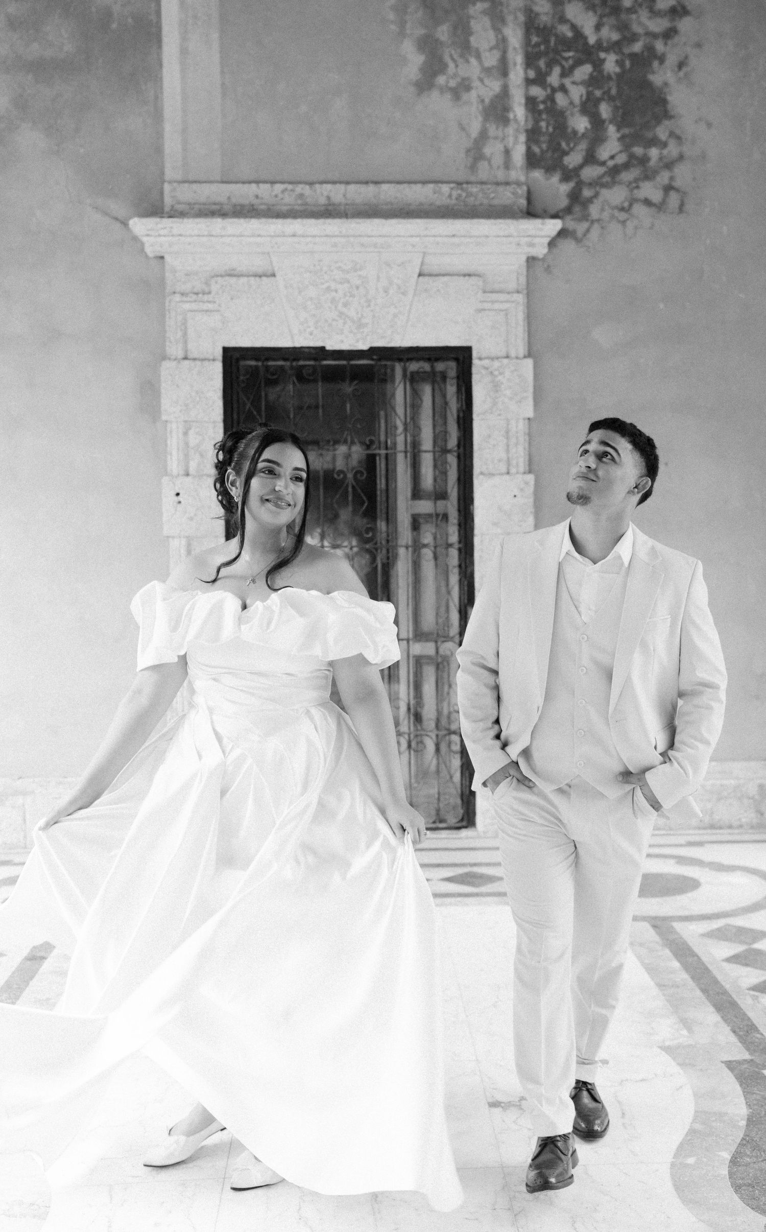 Two people in white formal attire dance gracefully in an artistic black and white photograph against a building facade.