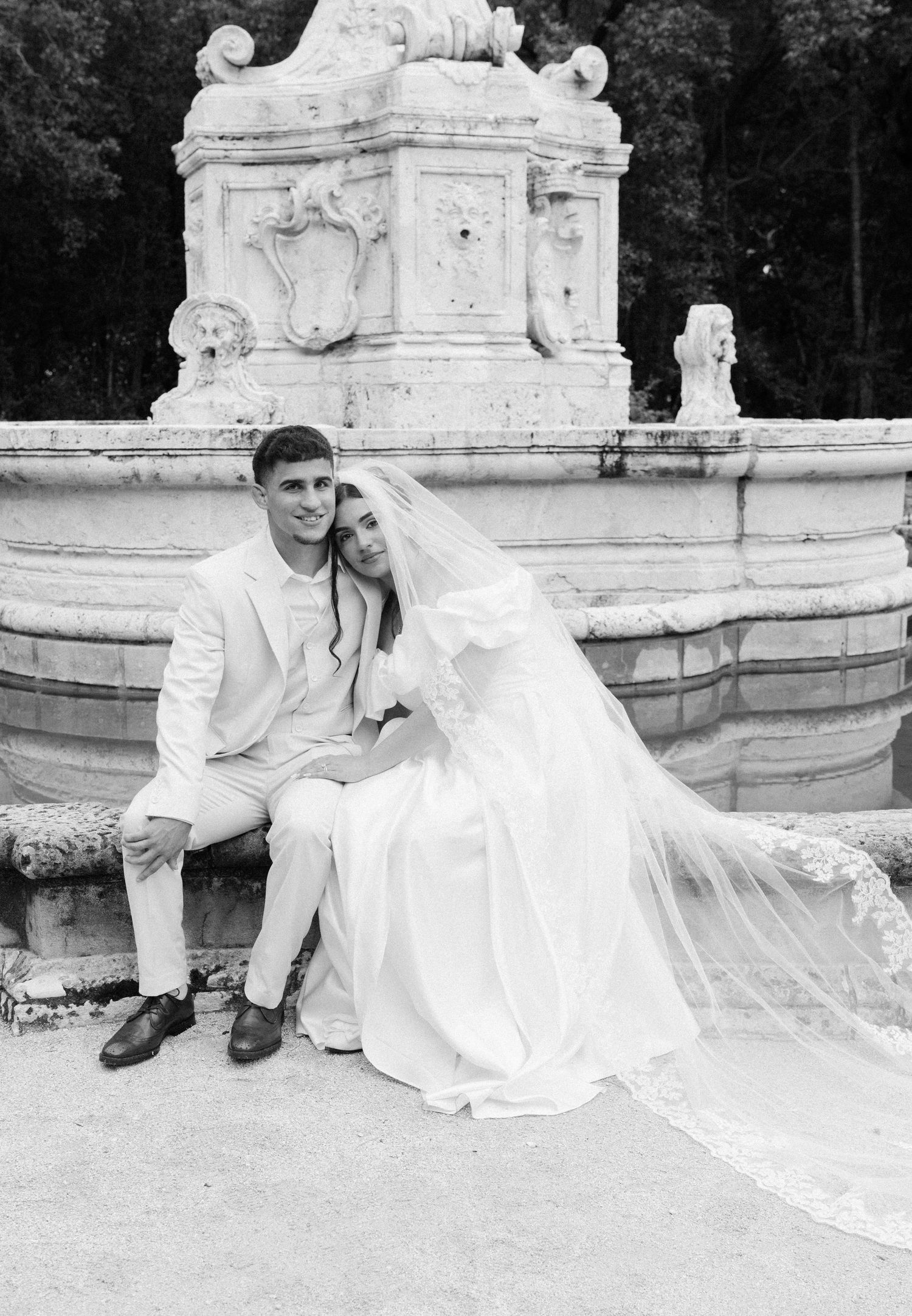 A black and white wedding photo of a couple sitting by an ornate stone fountain in a garden setting.
