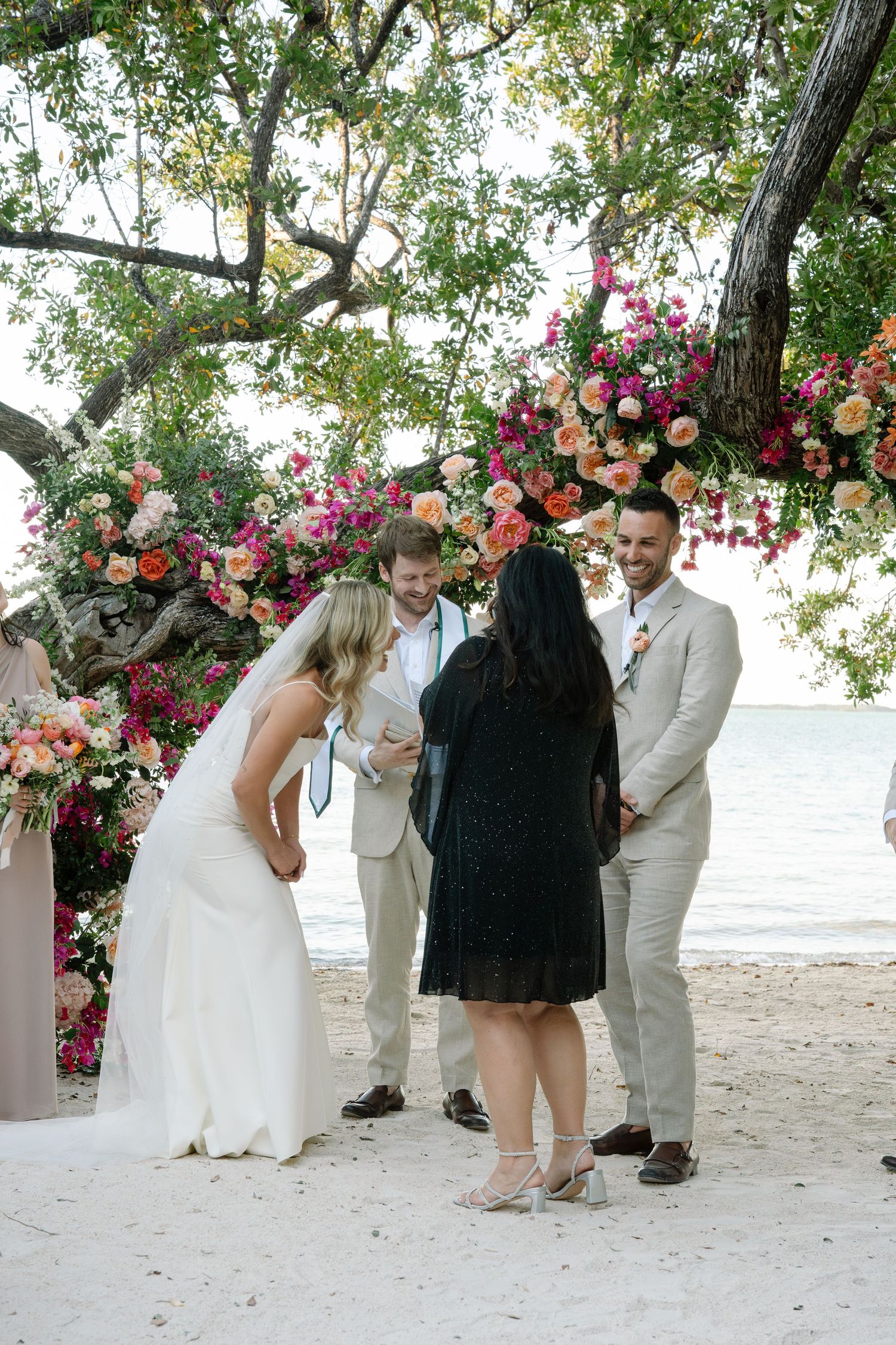 Wedding ceremony under a floral arch with pink and coral blooms on a beach.