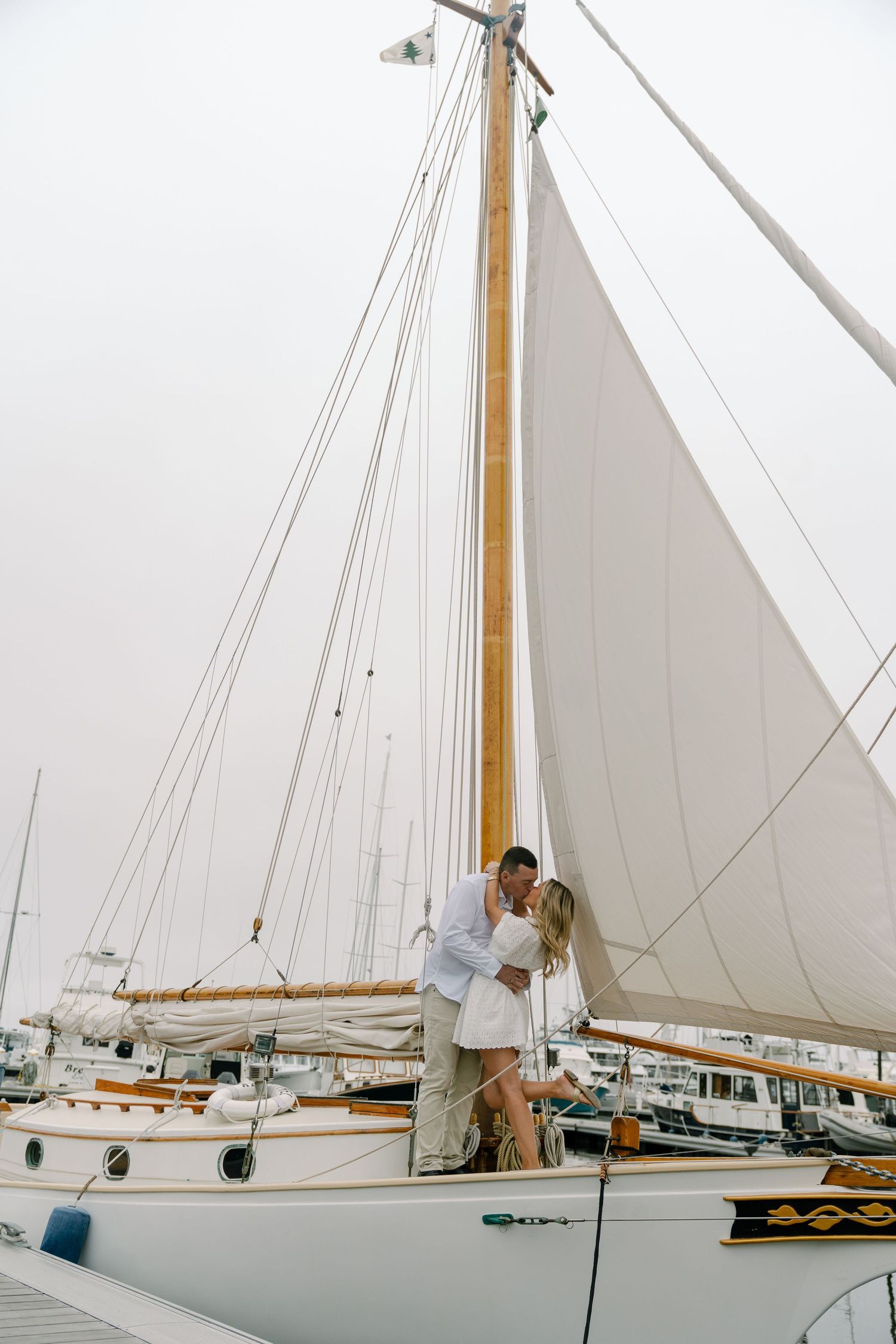 Couple sharing a romantic kiss on a classic white sailboat at a marina, surrounded by tall masts and rigging.