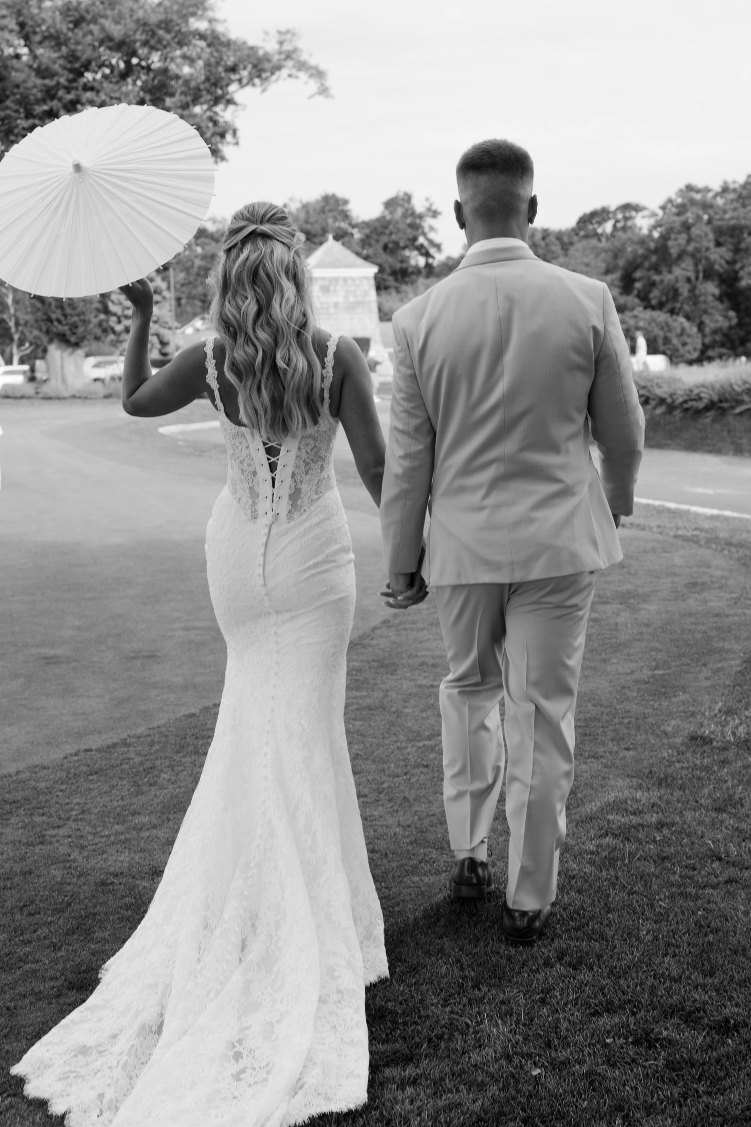 Black and white photo of bride in lace gown holding parasol walking hand in hand with groom on green lawn.