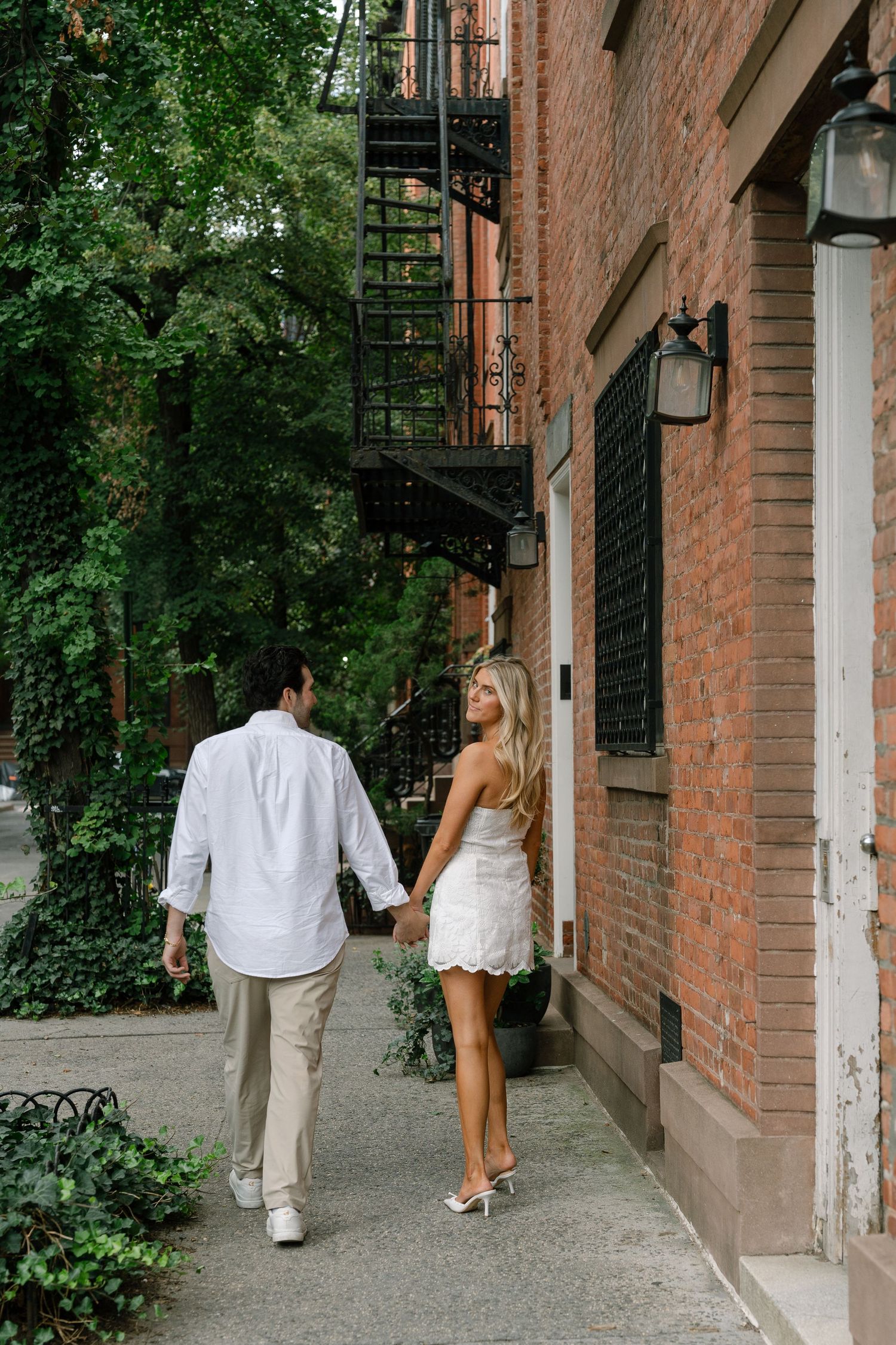 Couple holding hands walking along a brick building sidewalk, woman in white mini dress looks back, lush greenery in background.