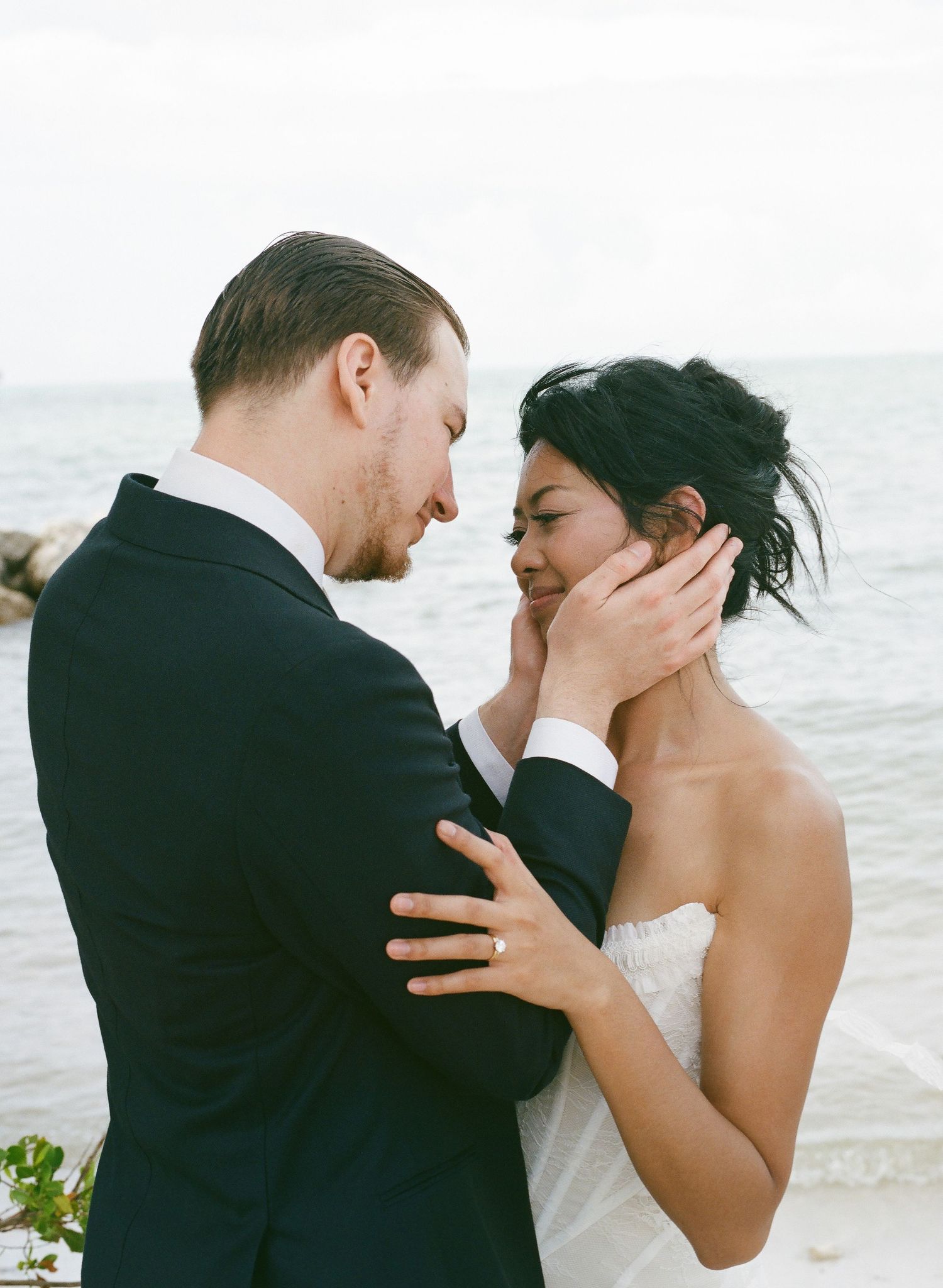 Romantic couple sharing an intimate moment on a beach, groom in black suit tenderly holding bride's face near the ocean.