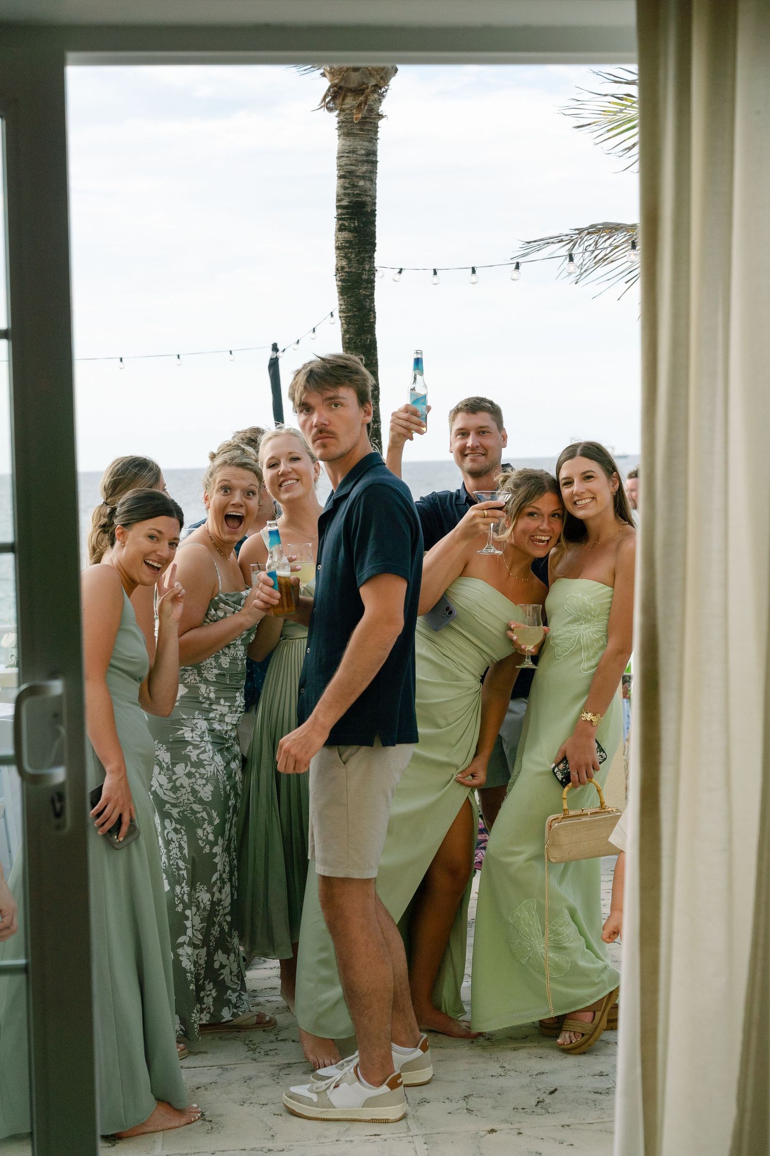Group of friends in sage green dresses celebrating at a beach wedding, raising drinks and smiling by a palm tree.