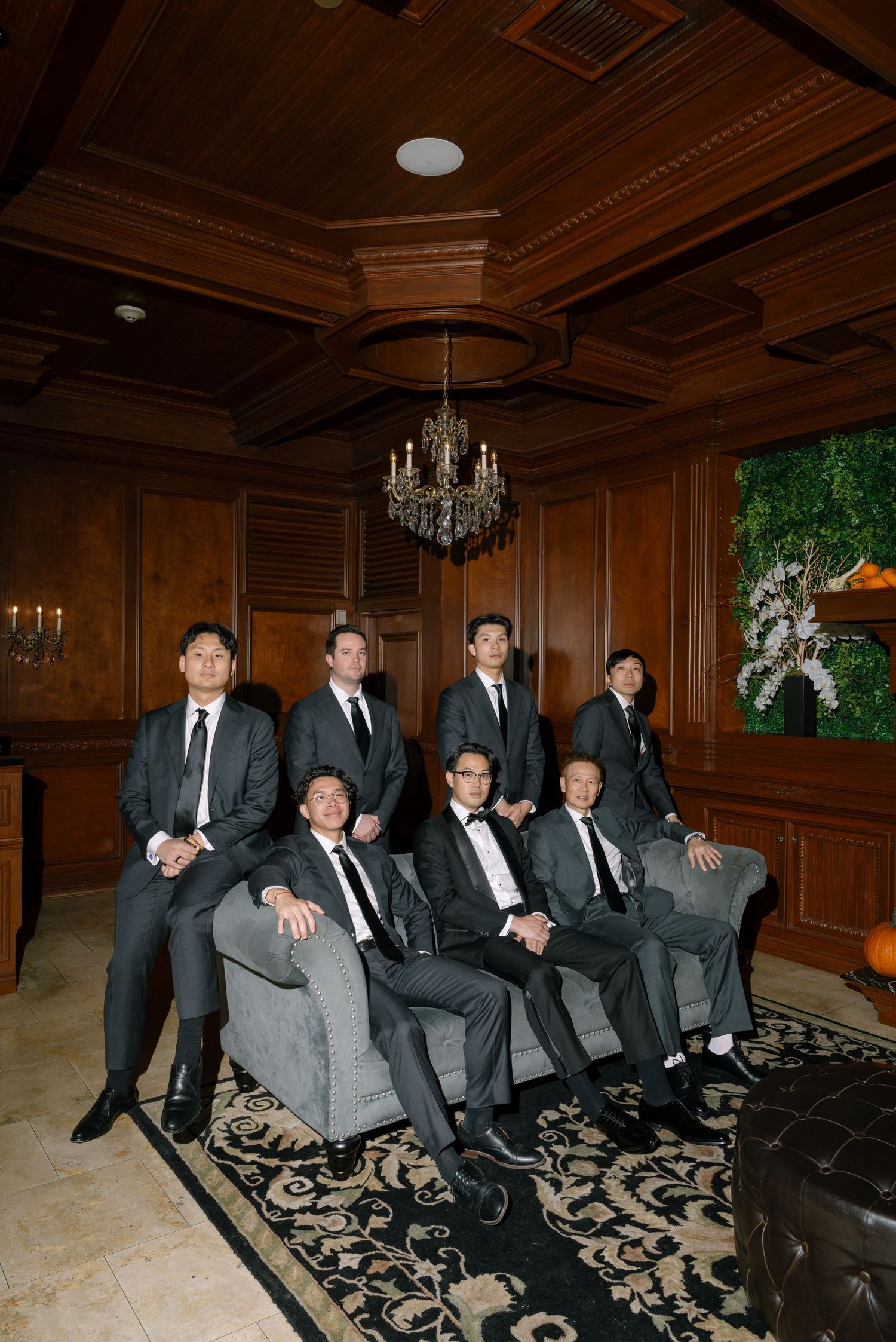 Groomsmen in black suits and ties lounging on elegant furniture in a wood-paneled room with chandeliers.