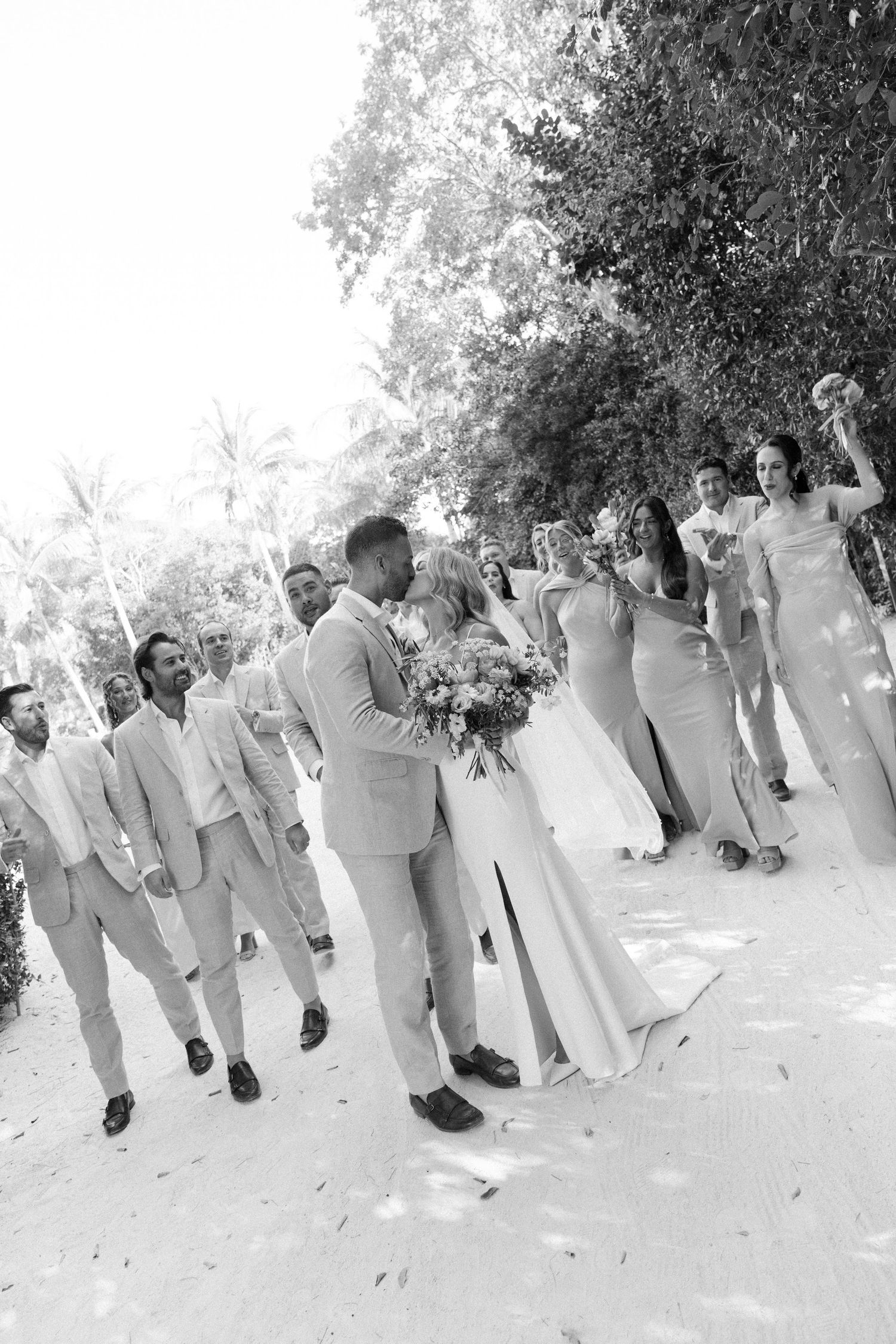 Wedding party celebrates on a sunlit path surrounded by lush greenery.
