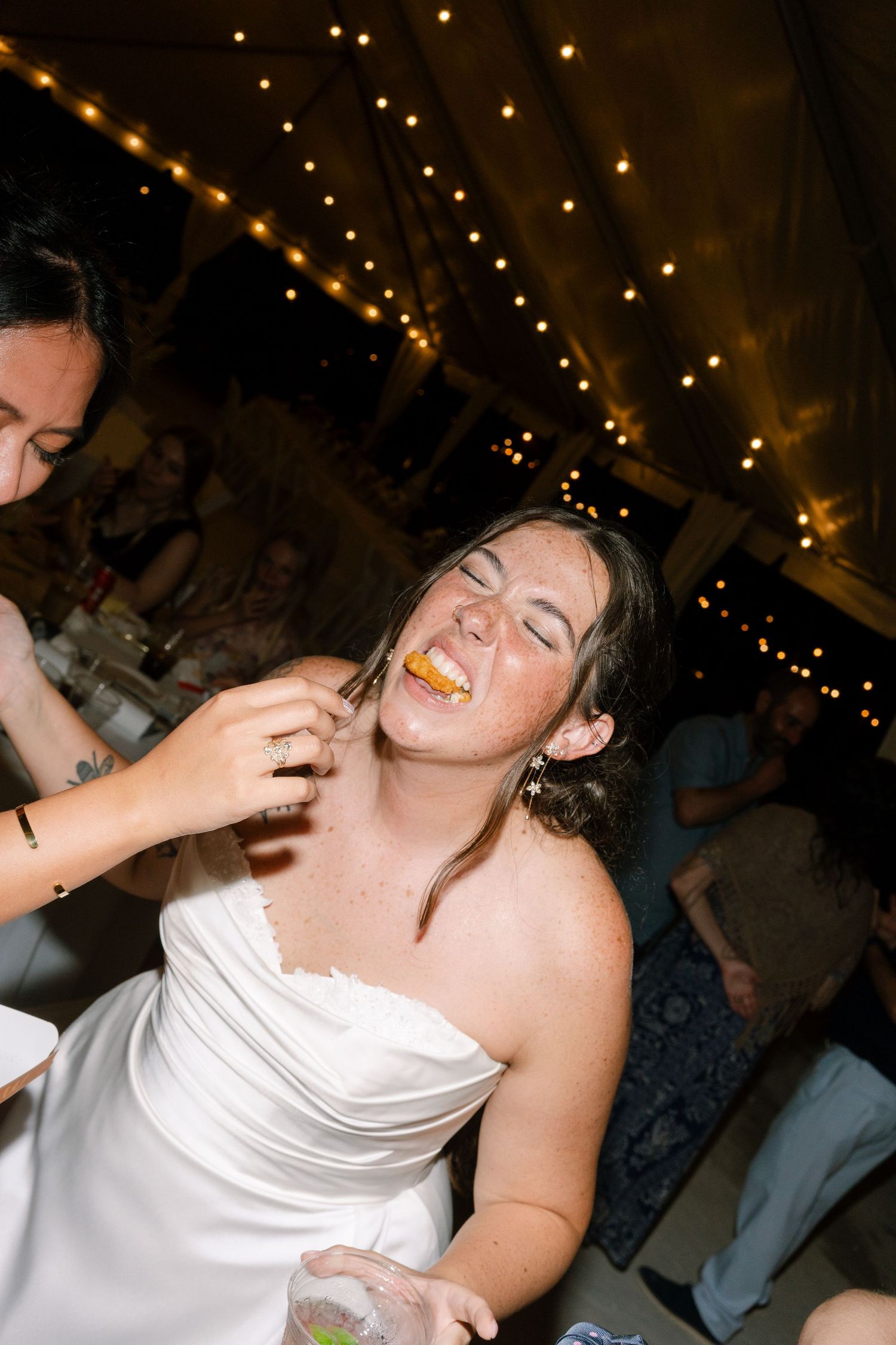 A woman in a white dress smiling and having her makeup applied by another person.