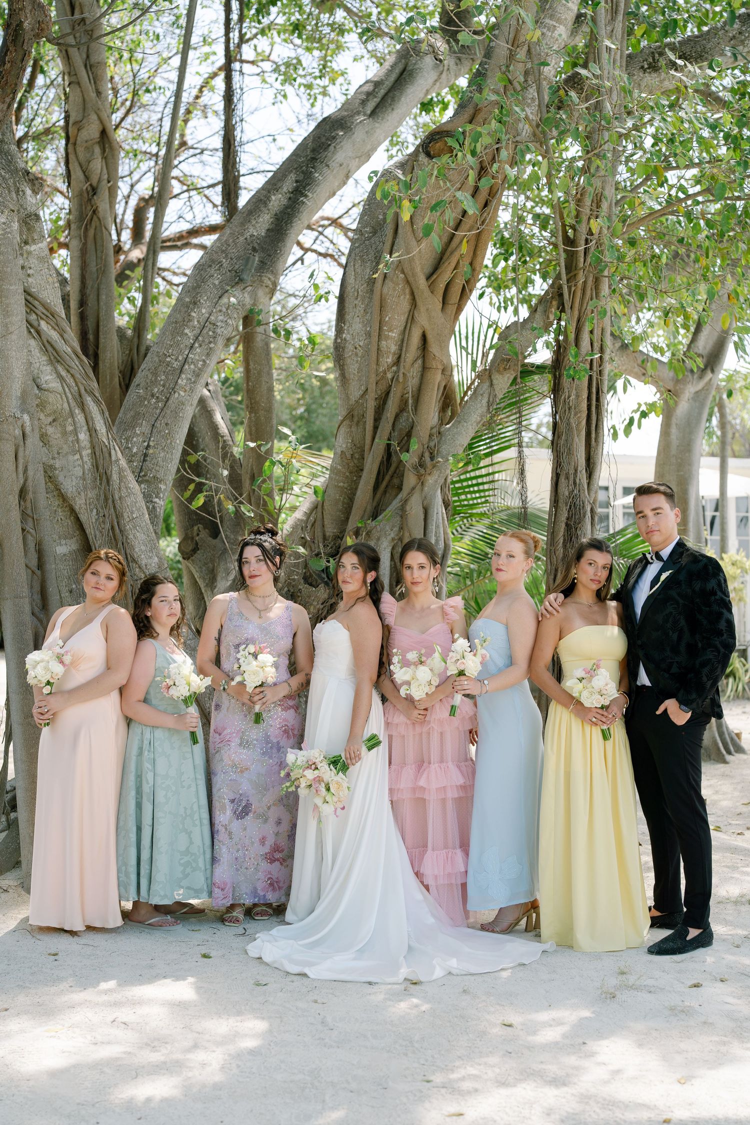 A group of people in formal attire, including a bride and groom, stand in a lush, green outdoor setting.