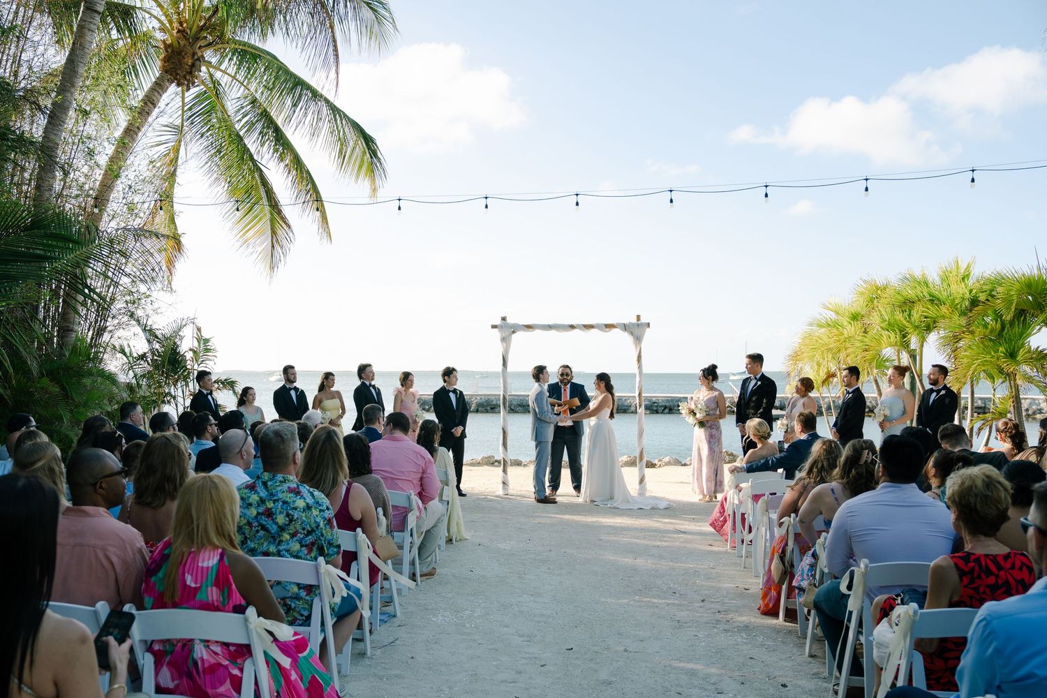 A scenic beach wedding ceremony with palm trees, guests, and an arch.