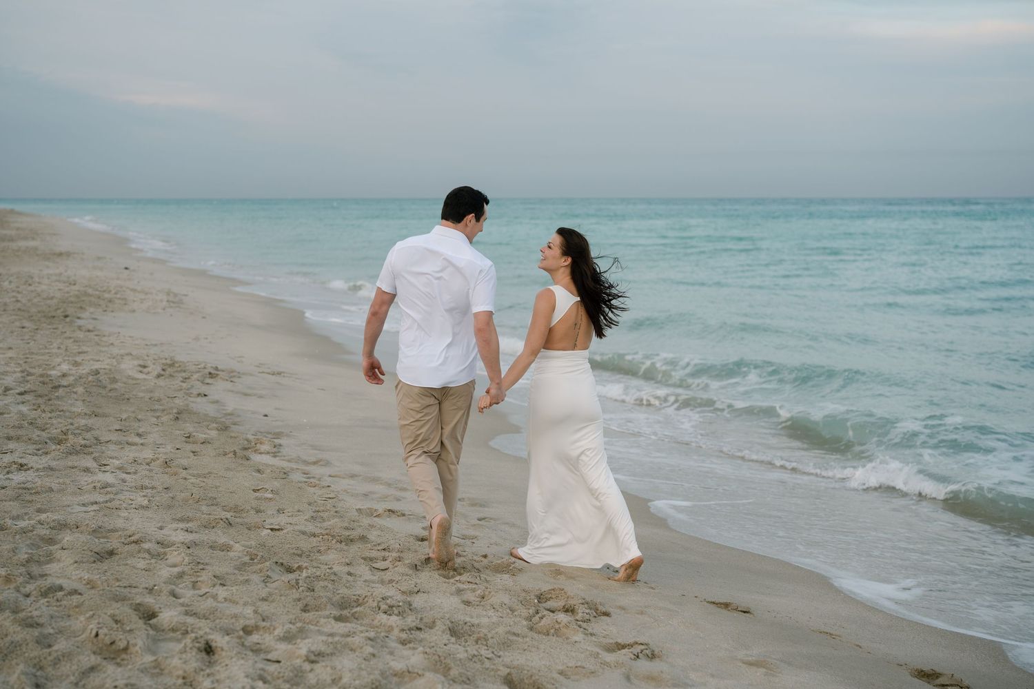 A romantic couple walks hand in hand along a serene beach with turquoise waters and soft sand at sunset.