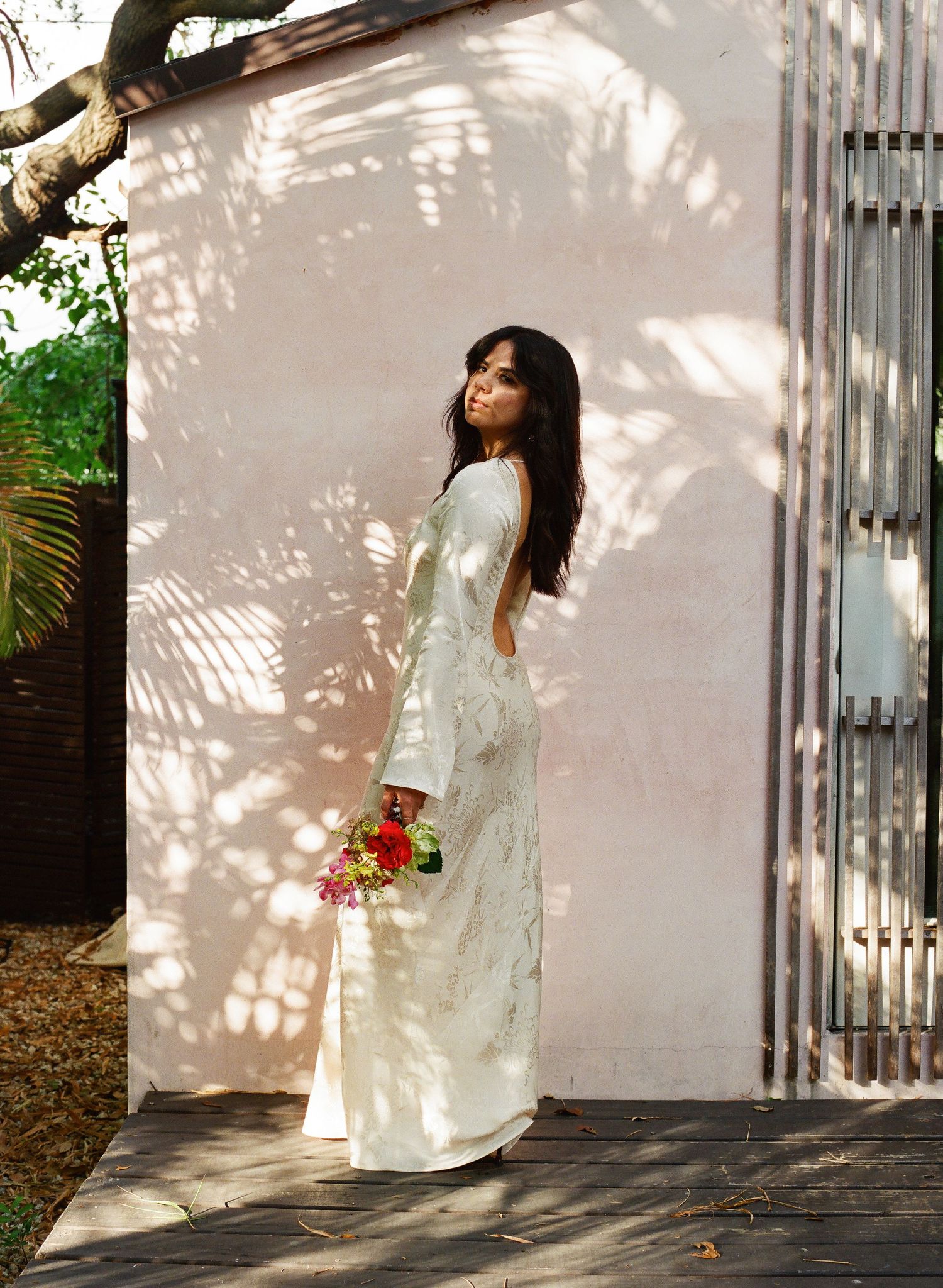 Person in white dress poses against sunlit wall with dappled shadows.