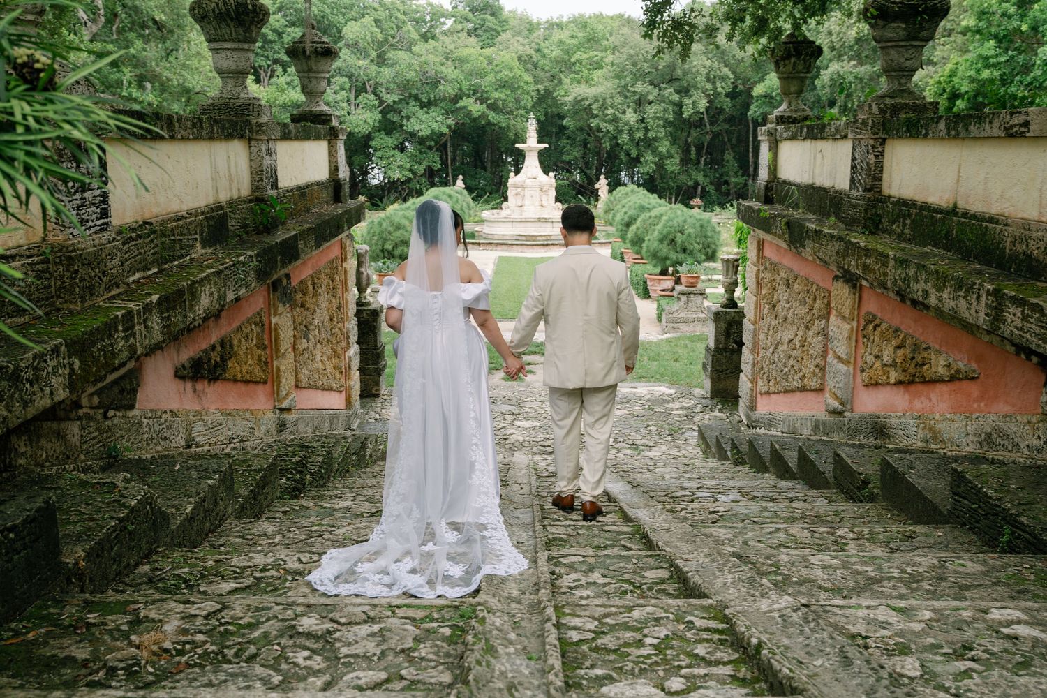 A couple in wedding attire walks down a rustic stone path lined with brick walls and lush greenery.
