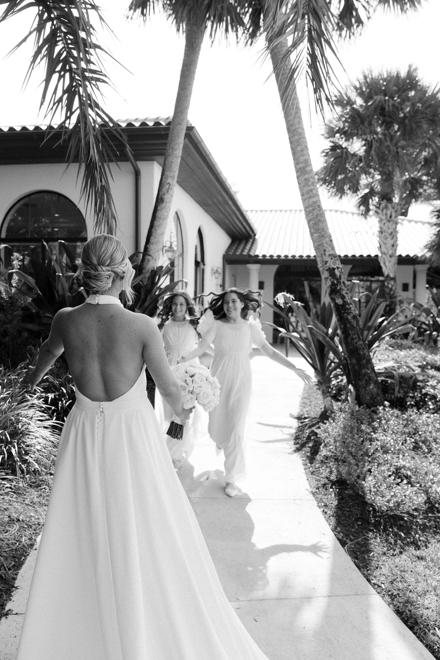 Black and white photo of people in white dresses walking along a palm tree-lined path outside a Spanish-style building.