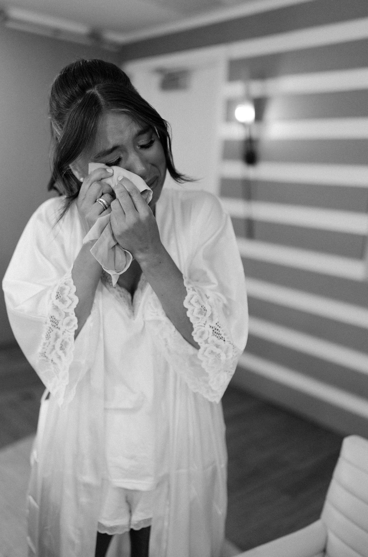 A person in a white lace robe wipes tears with a tissue in a black and white emotional moment.