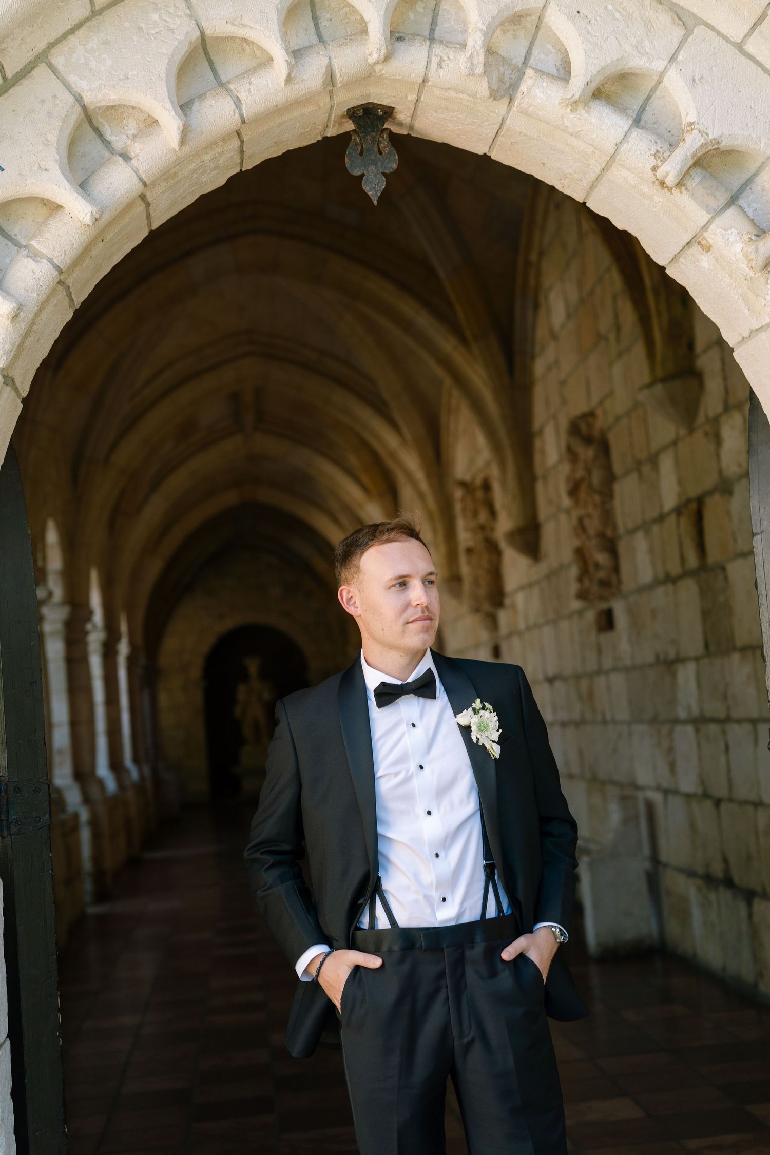 A groom in a black tuxedo with bow tie and boutonniere stands in a stone arched corridor.