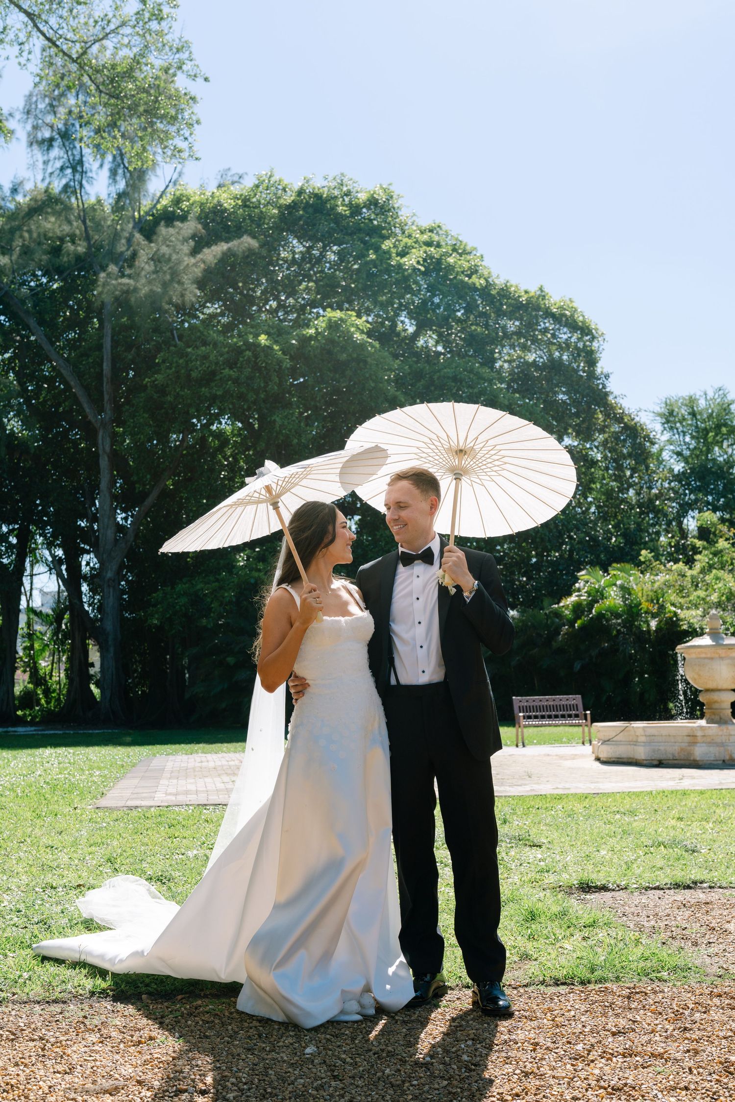 Bride and groom holding white paper parasols smiling at each other in sunny garden wedding venue.