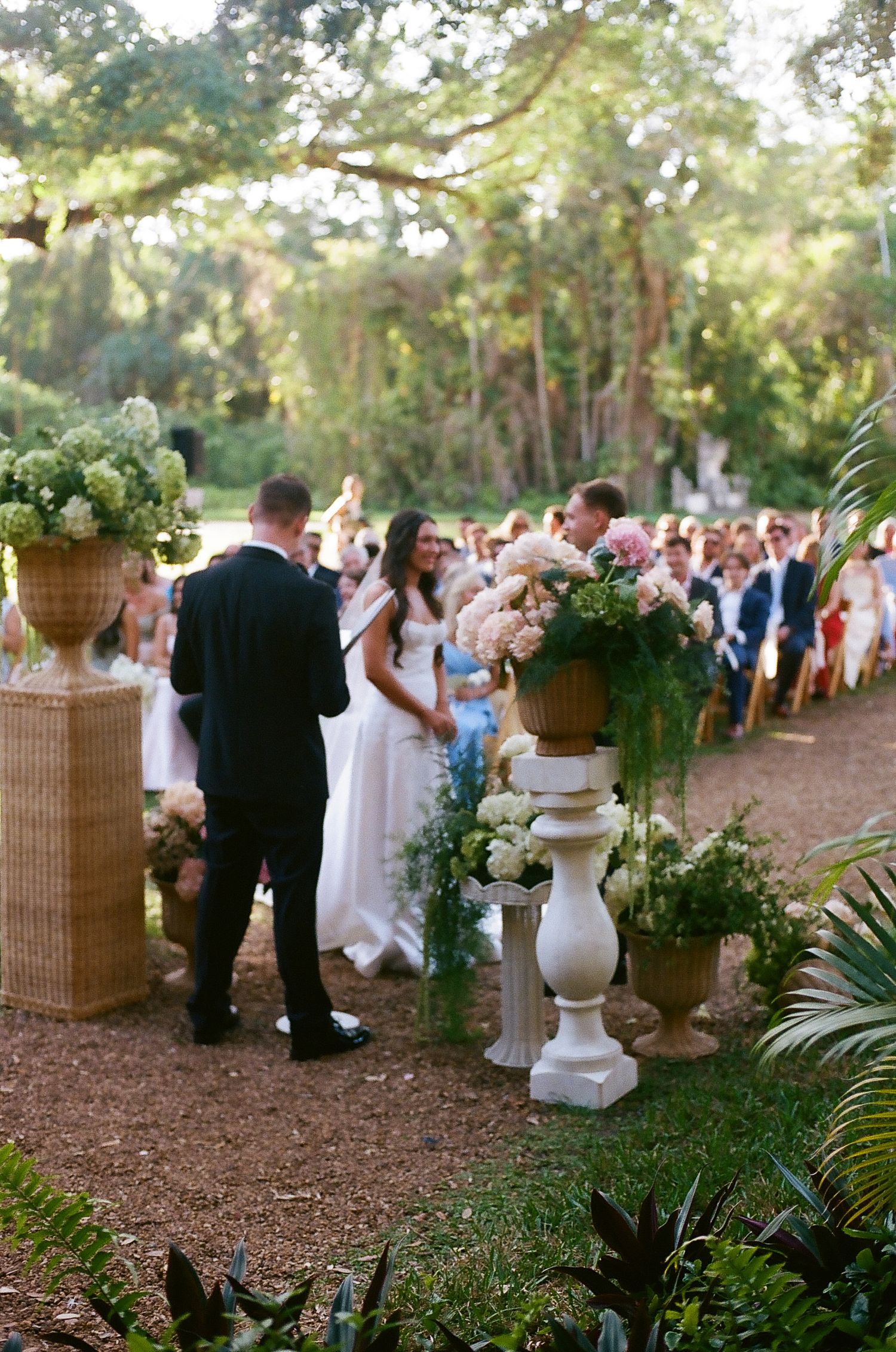 Outdoor garden wedding ceremony with couple at altar surrounded by lush floral arrangements in urns.