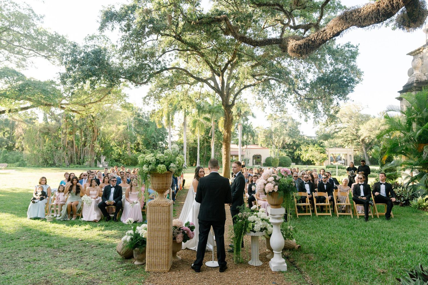 Outdoor wedding ceremony under large oak tree with guests seated on both sides in lush garden setting.