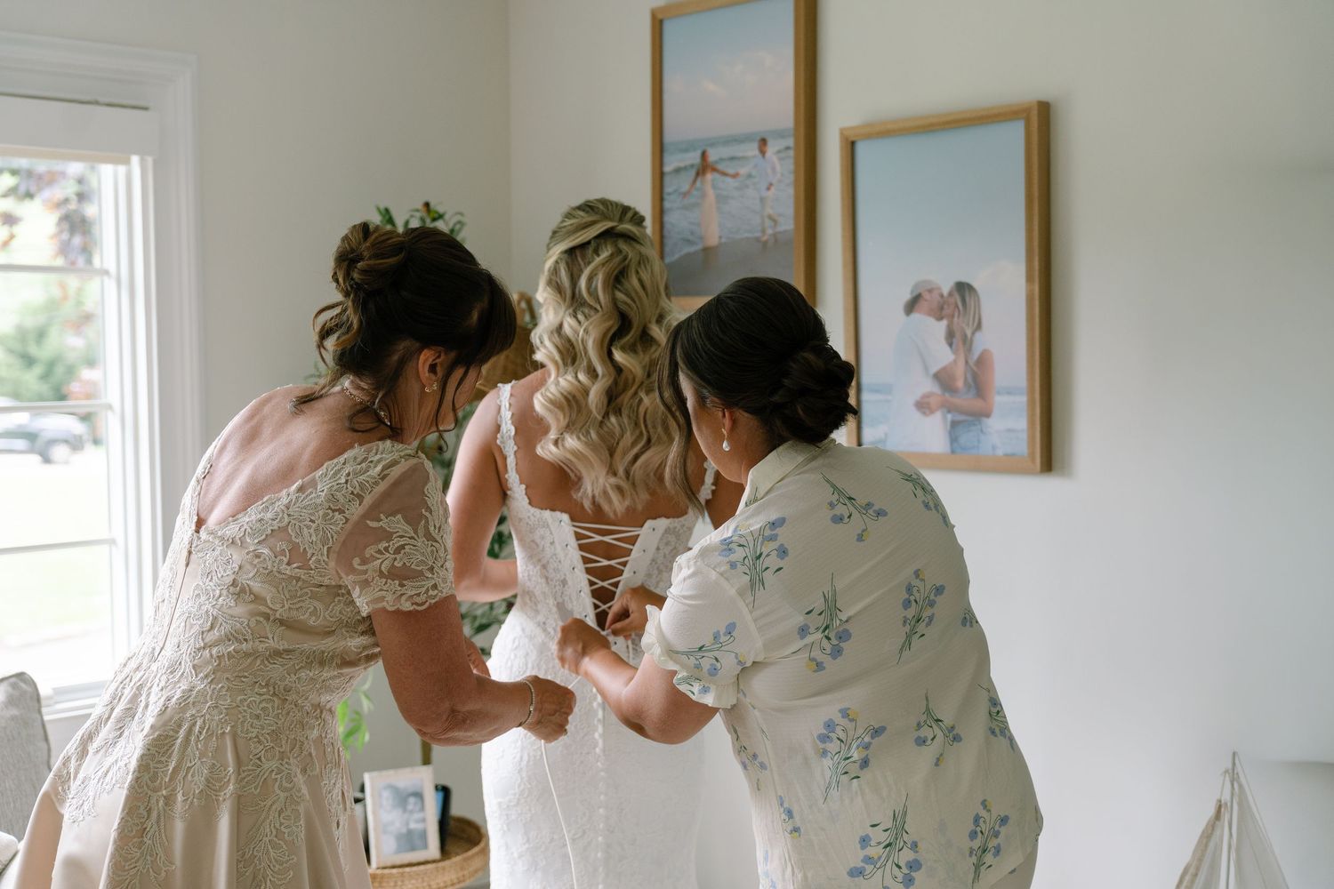 Two bridesmaids help a blonde bride lace up her white wedding dress indoors with coastal artwork on the wall.