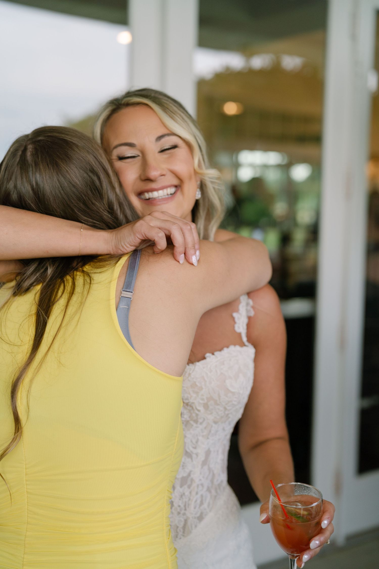 A smiling bride in a lace gown hugs a guest in a yellow dress while holding a cocktail at her wedding reception.