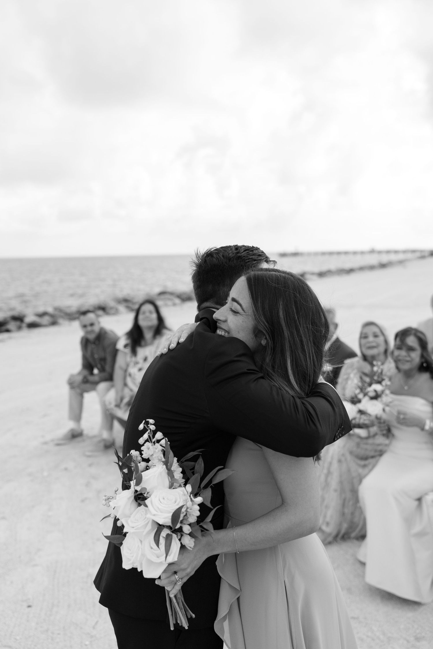 Black and white photo of a bride and groom embracing on a beach during their wedding ceremony.