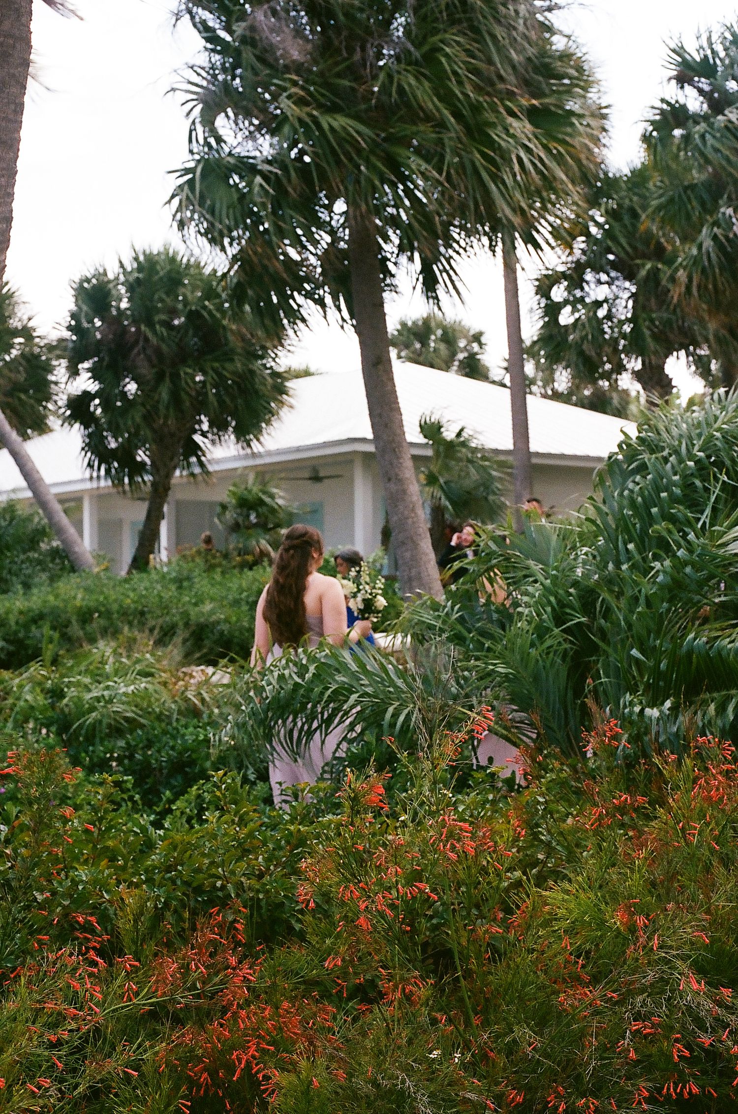 Bridesmaid in floral dress holding white bouquet walking through lush tropical garden near white venue building.