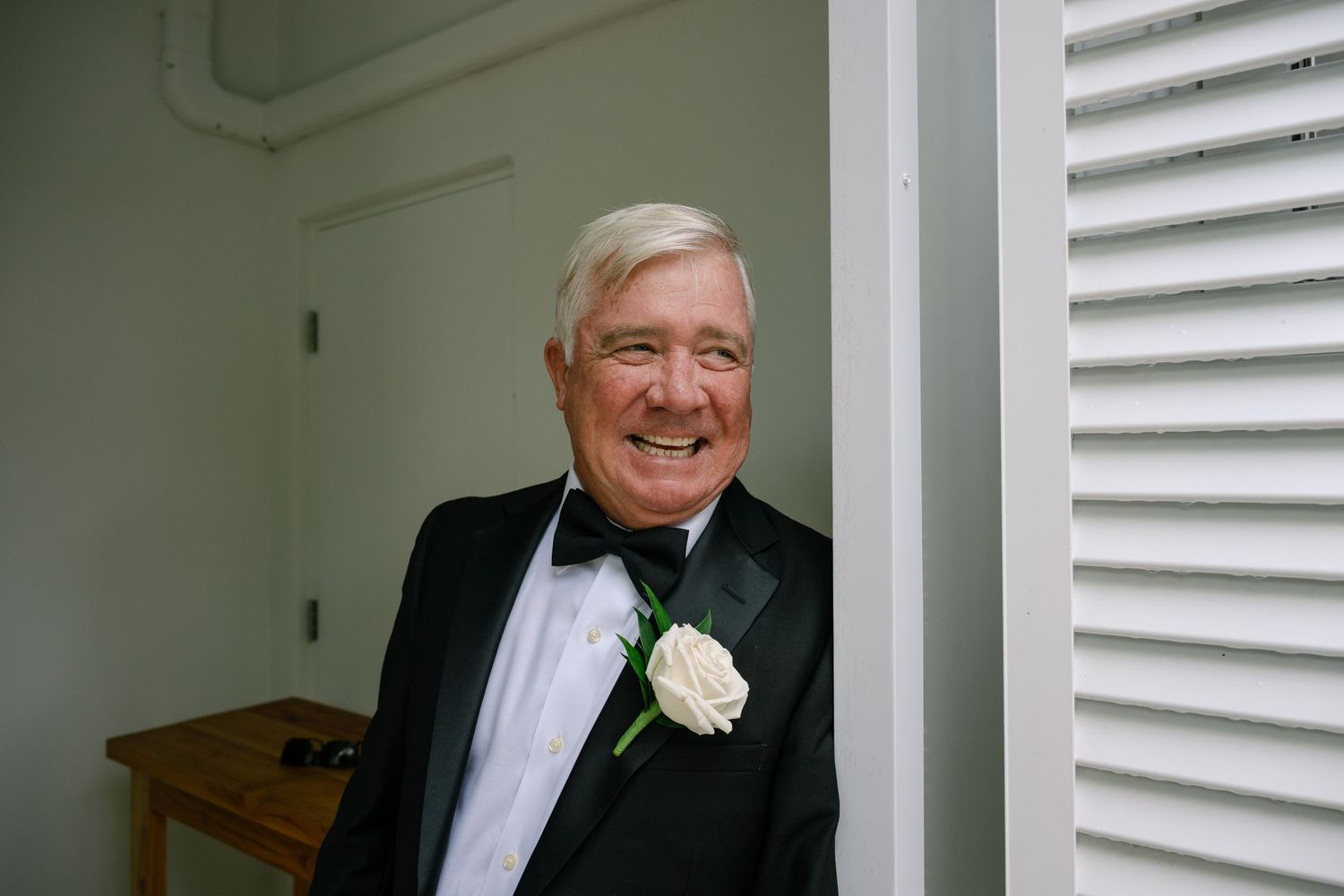 Smiling older gentleman in black tuxedo with white rose boutonniere at wedding celebration.
