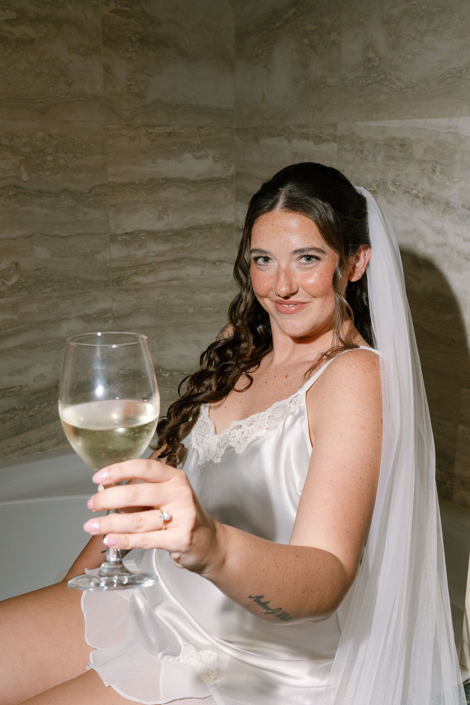 A smiling bride in a white wedding dress holds a glass of champagne while sitting on a couch.