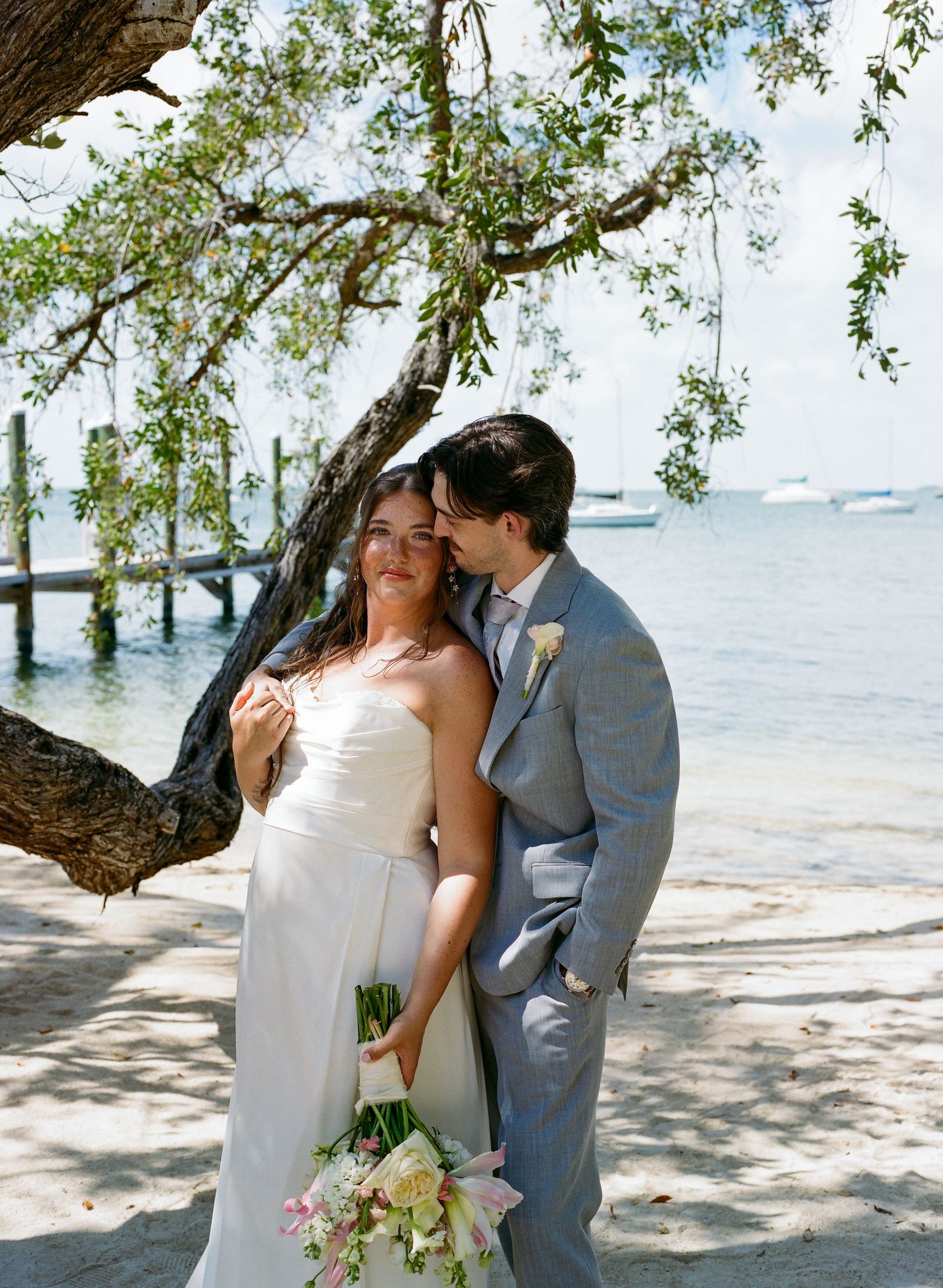 Bride and groom share a tender moment under a tree by the waterfront on their beach wedding day.