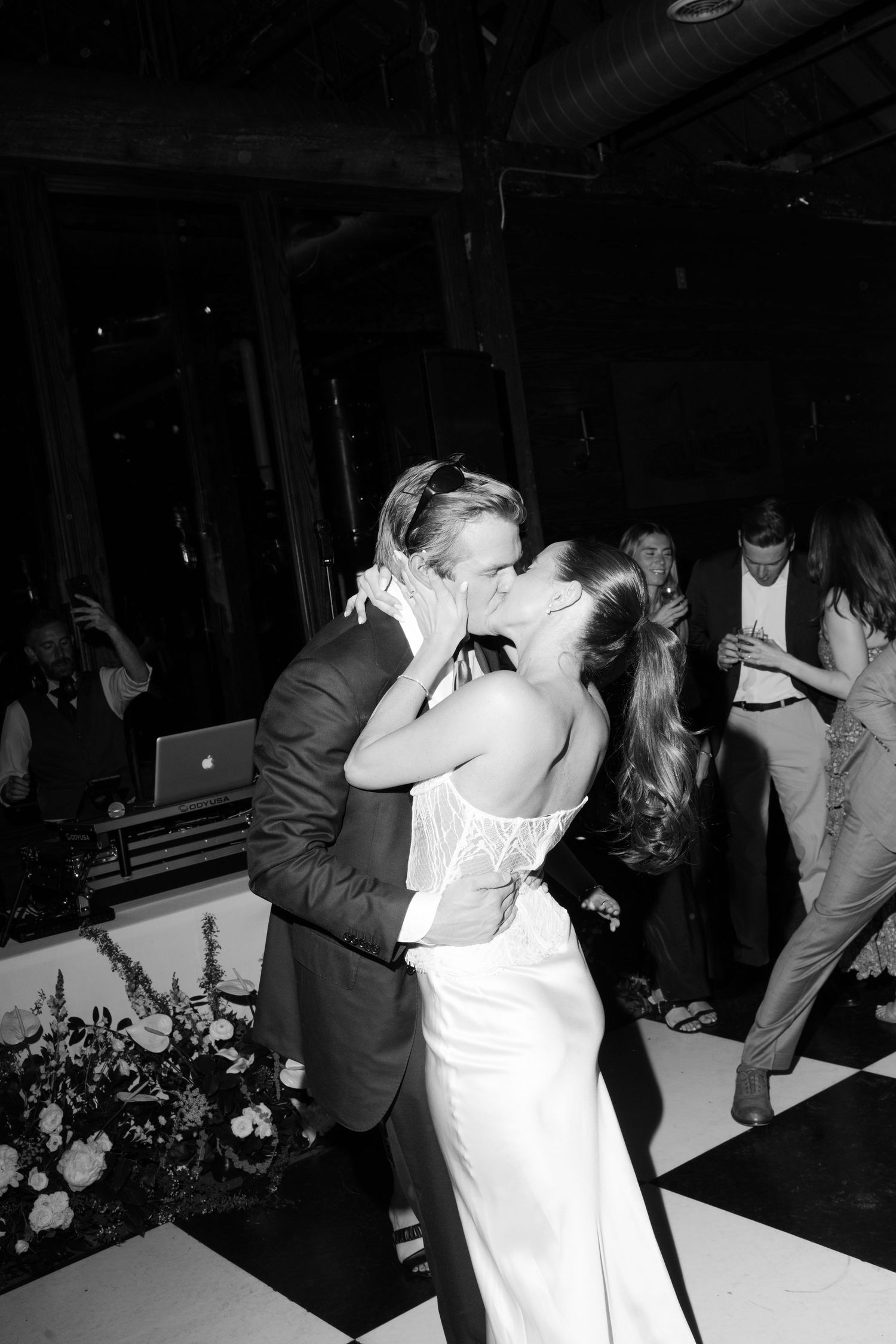 Black and white photo of a couple sharing a first dance kiss at their wedding reception on a checkered dance floor.