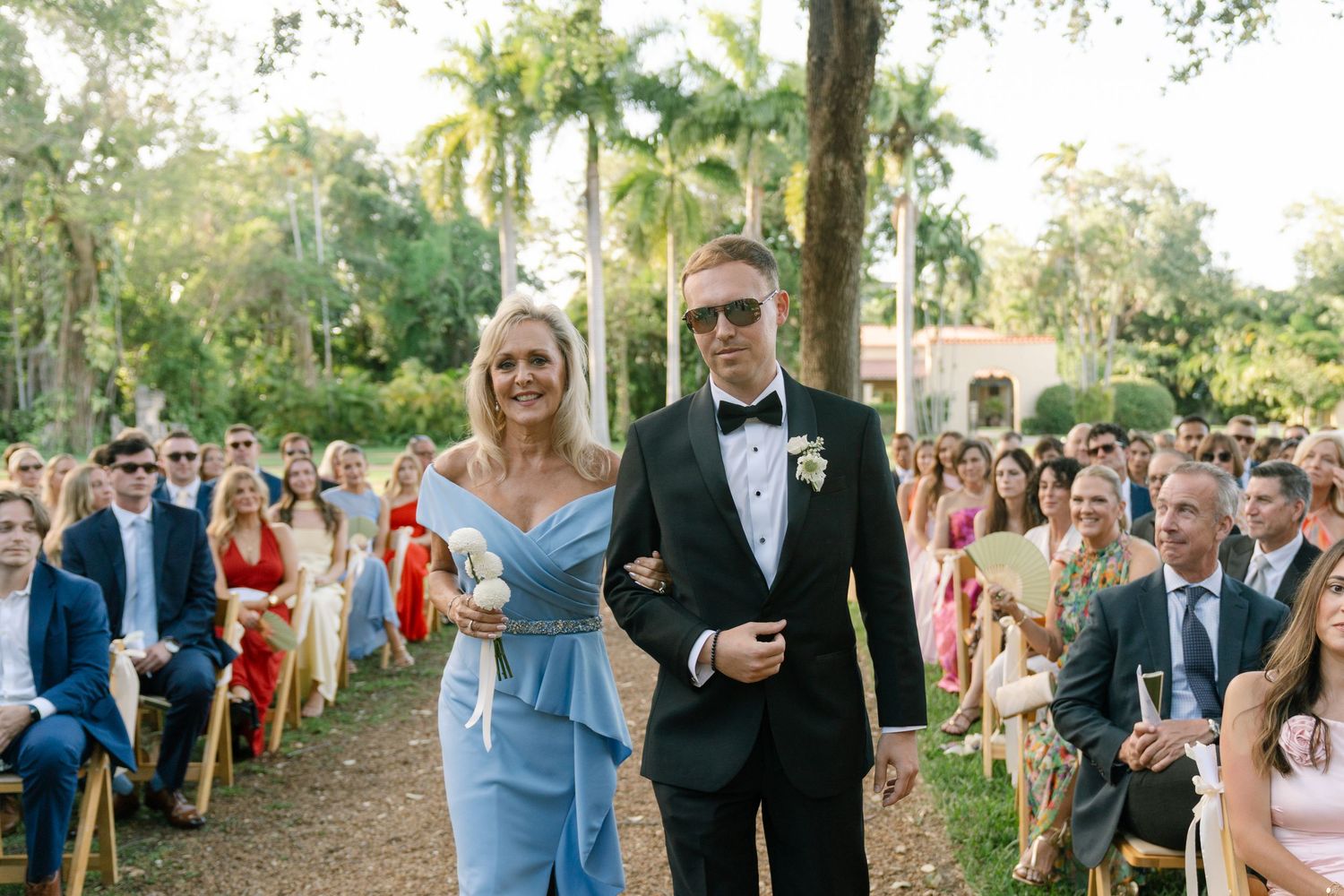 Groom in black tuxedo walks down outdoor wedding aisle with blonde woman in light blue off-shoulder gown.