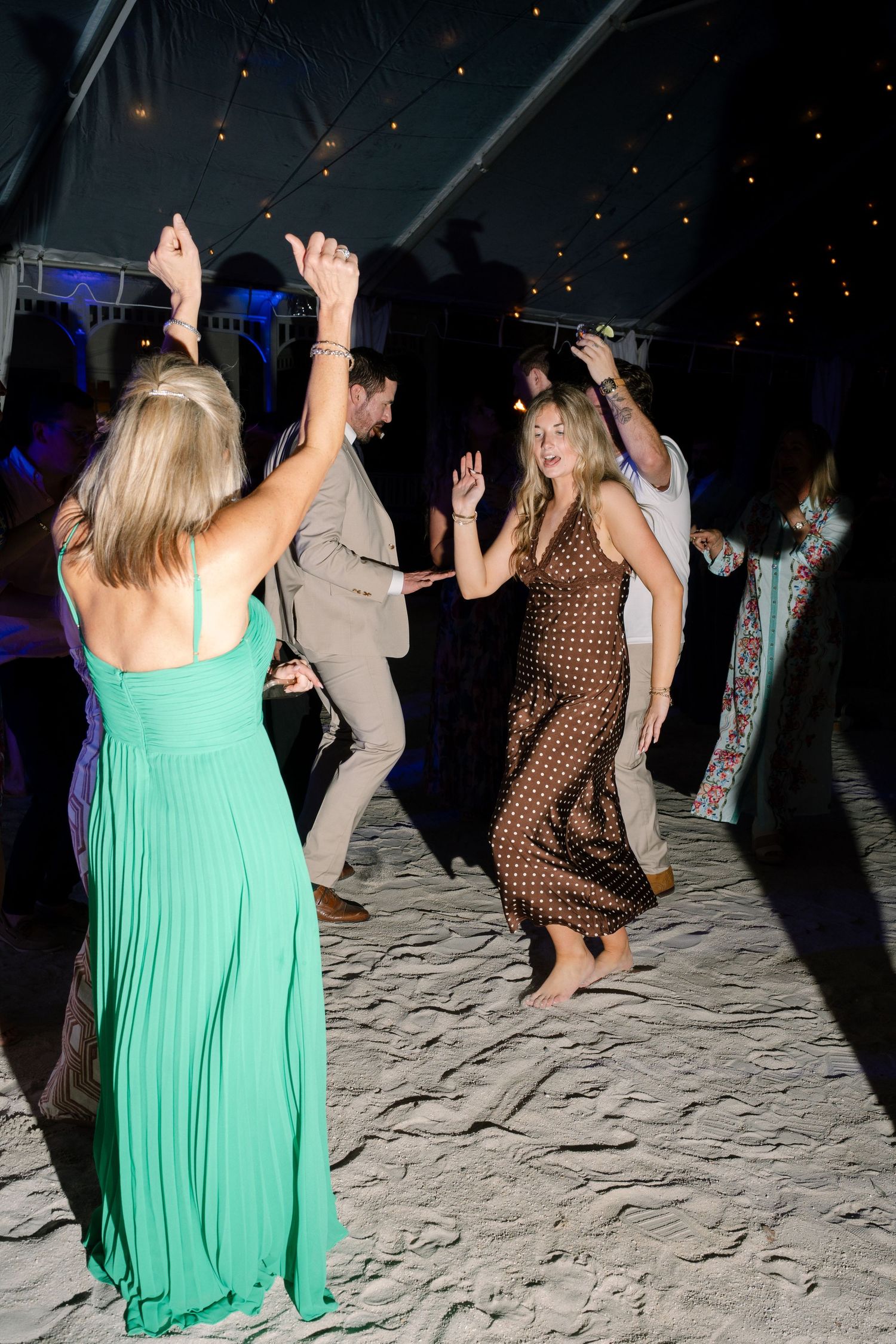Wedding guests dance and celebrate on a sandy floor under string lights at night.