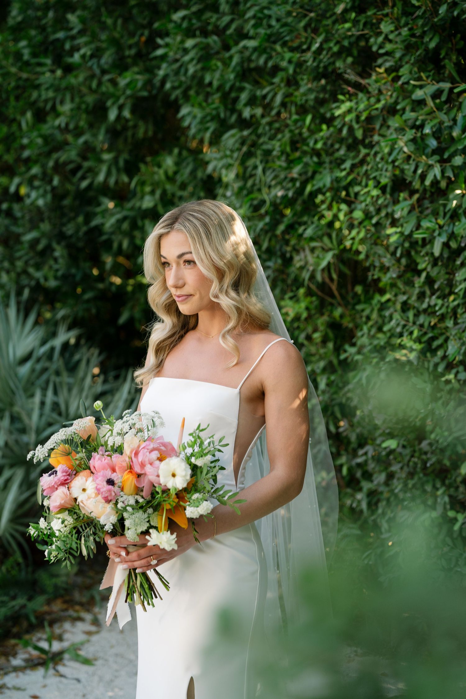 A bride in a white dress holds a vibrant bouquet of pink and orange flowers against a lush green garden backdrop.