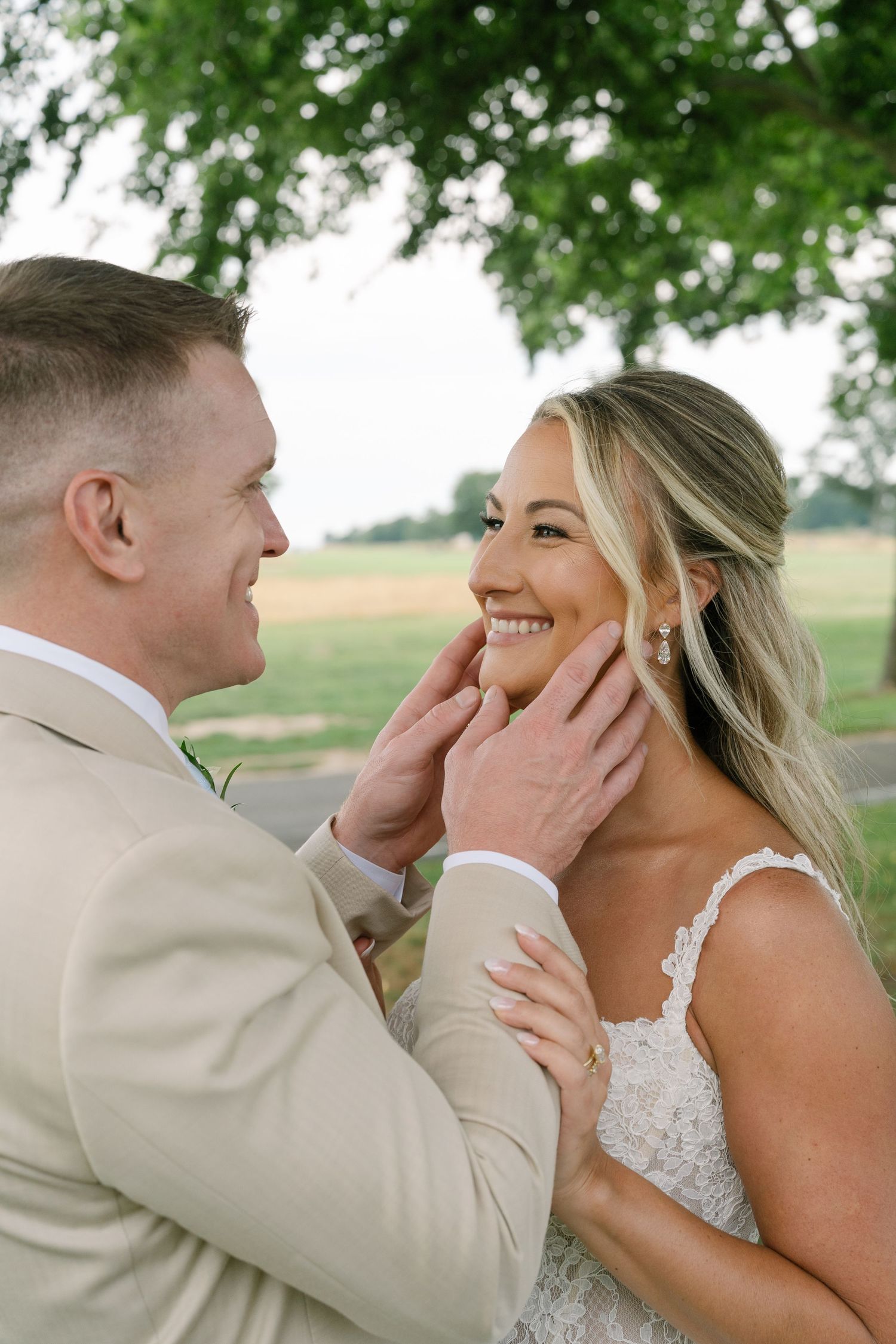 Groom in beige suit tenderly cupping smiling bride's face outdoors near green trees.
