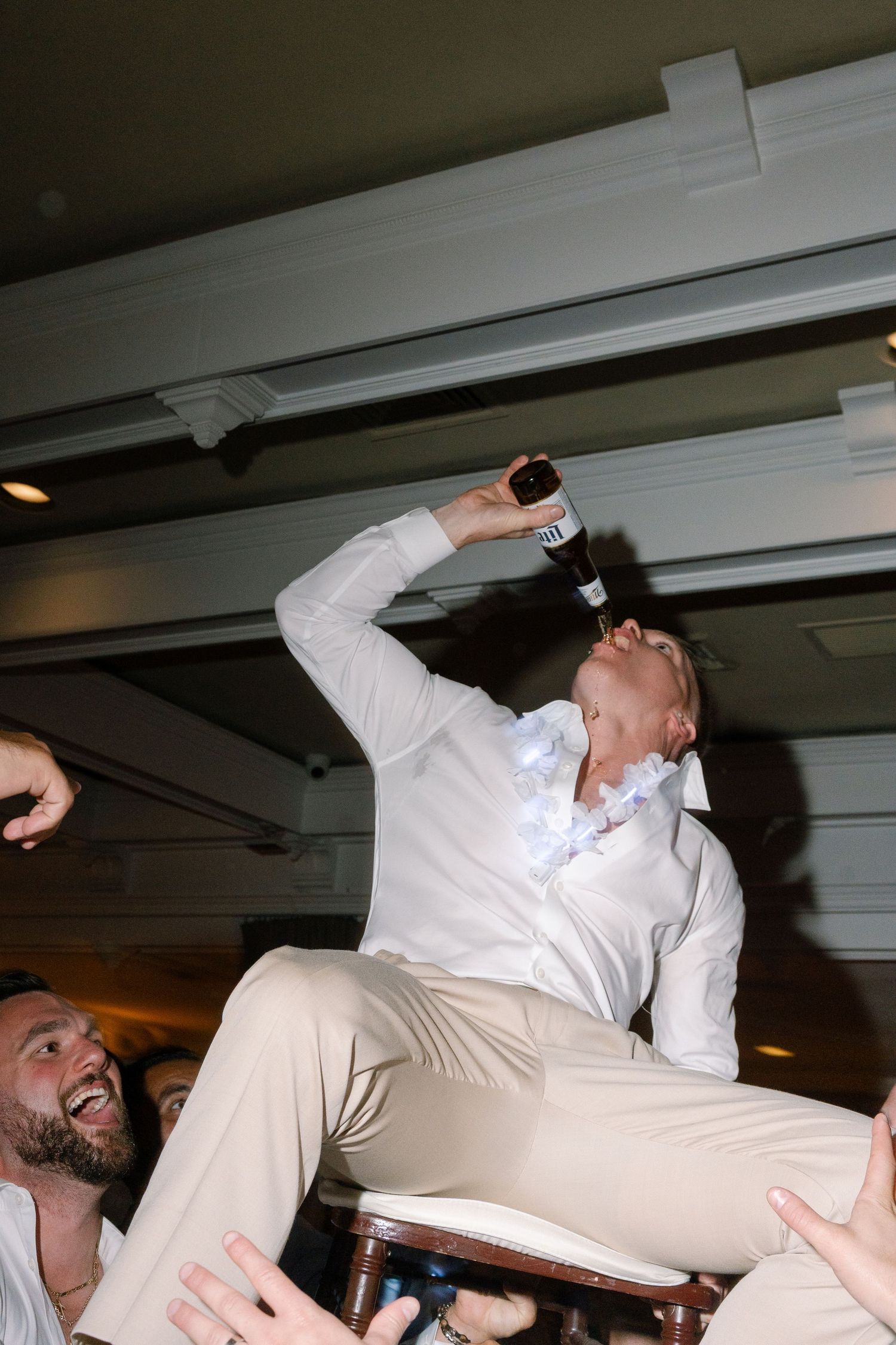 Wedding guest in white shirt drinking from bottle while being lifted on chair at reception.