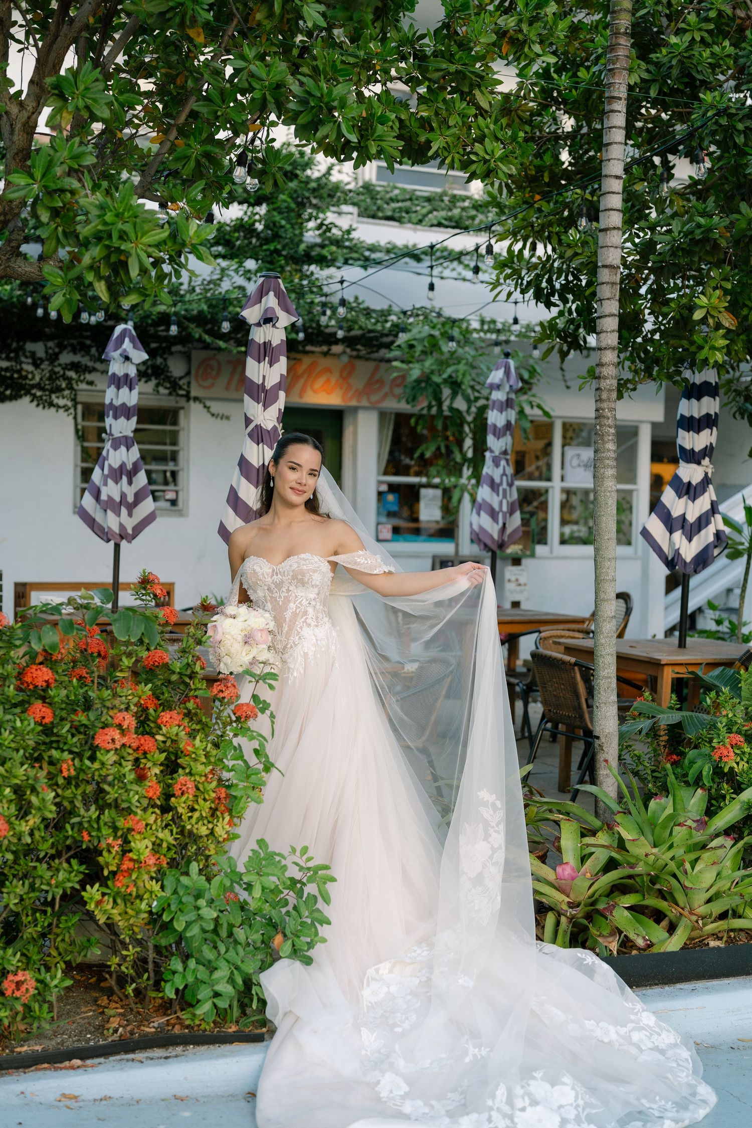 Bride in a lace ballgown holds her veil while posing outdoors among colorful flowers.