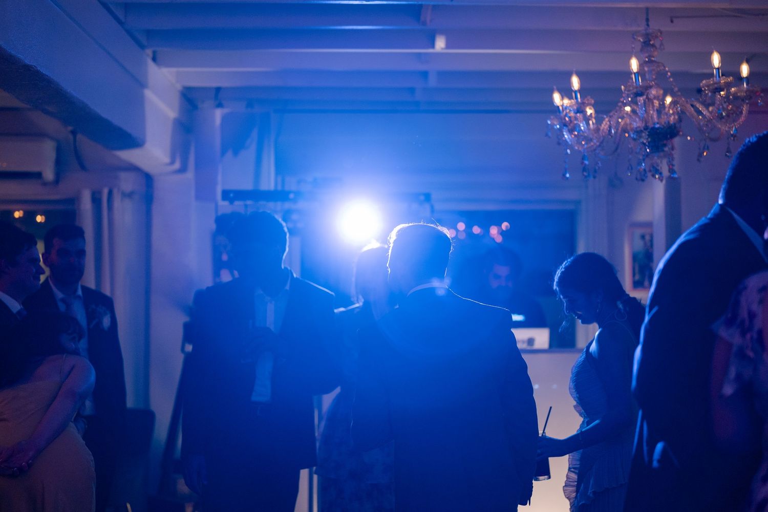 Elegant indoor party with guests silhouetted against bright stage lighting and a chandelier in a blue-lit venue.