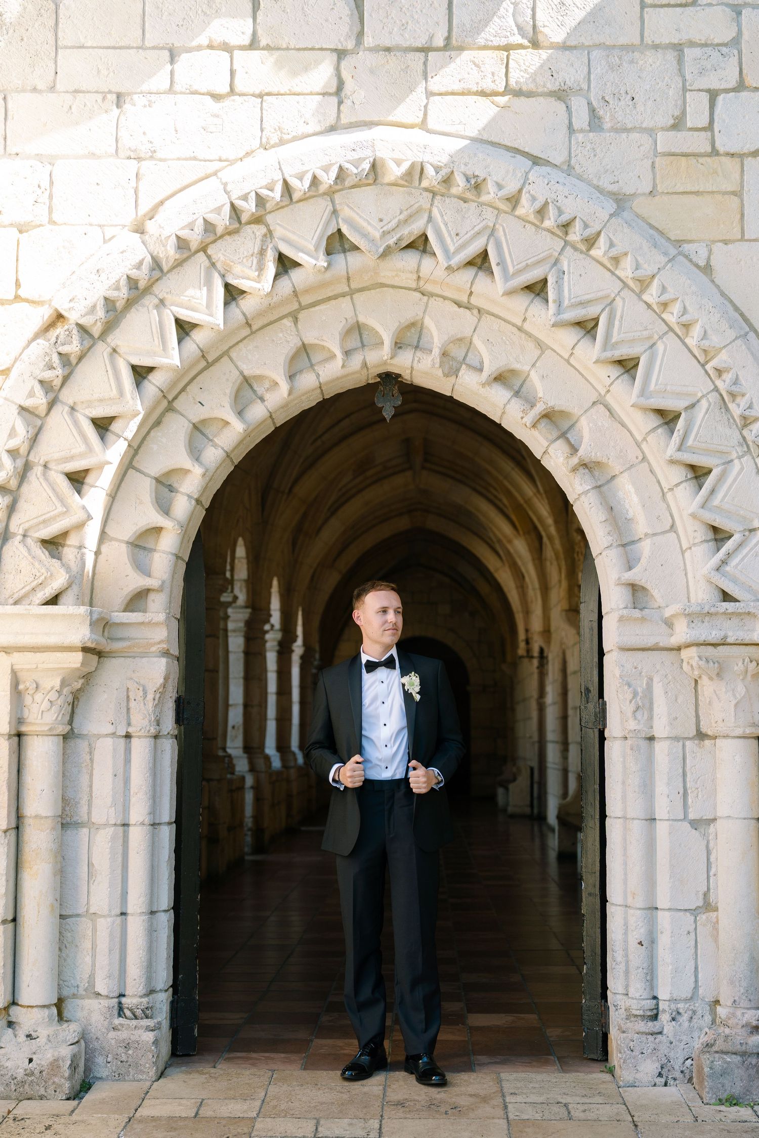 Groom in black tuxedo standing under ornate stone archway of historic building, elegant wedding portrait.