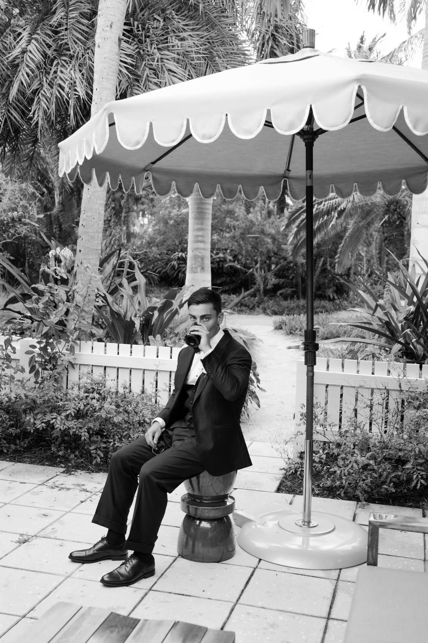 Black and white photo of a groom in a suit sitting under a striped umbrella surrounded by tropical palms.