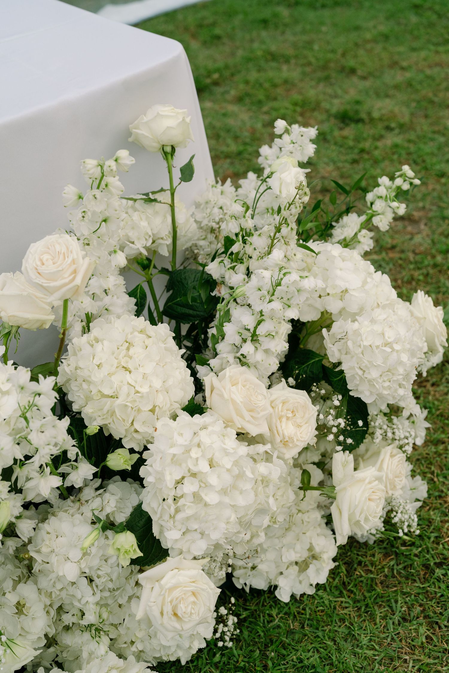 Lush white wedding floral arrangement featuring hydrangeas, roses, and delicate blooms on green grass.